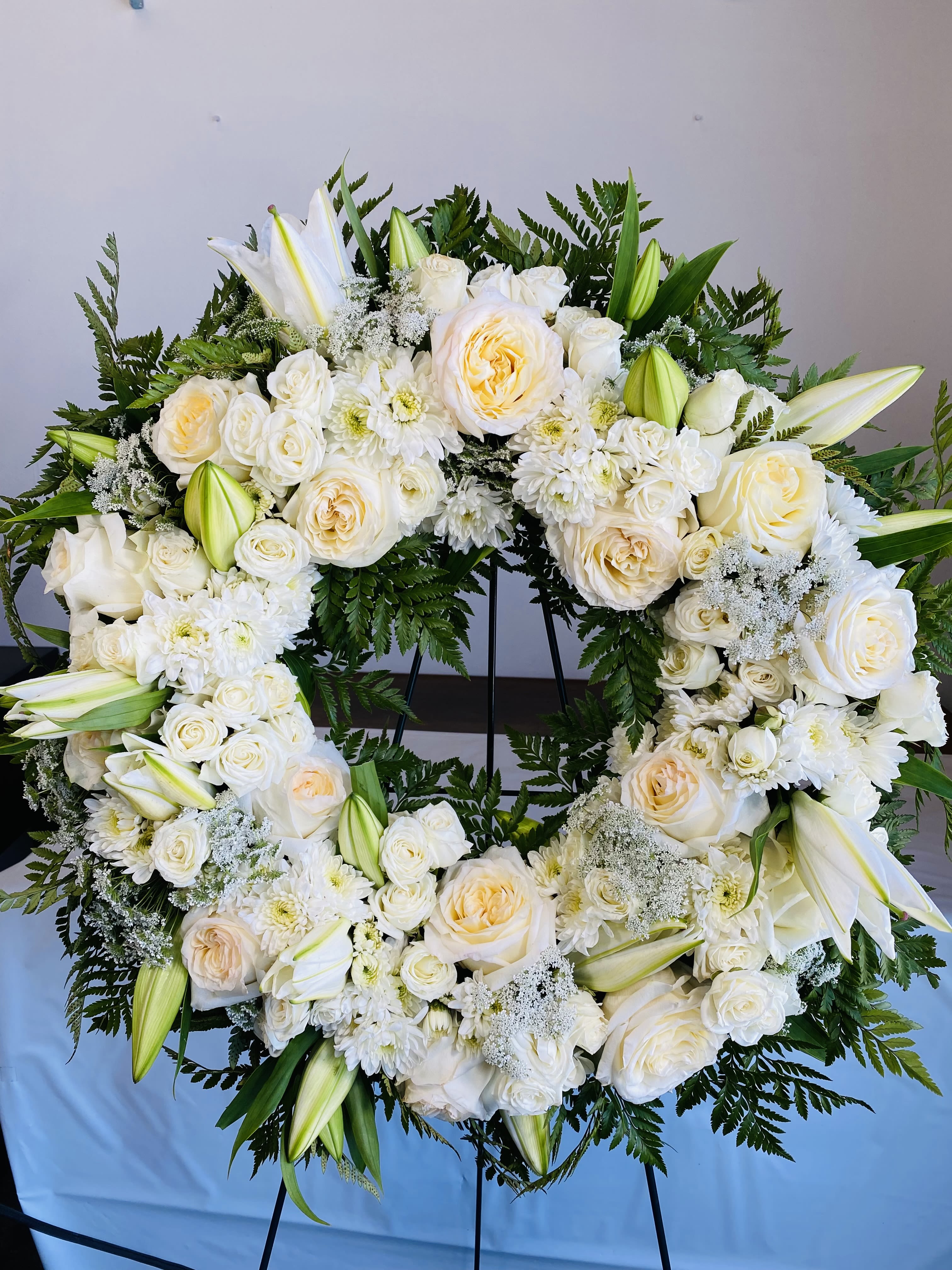 Elegant Ceremonial - The Elegant Ceremonial standing spray is a graceful and timeless floral tribute designed to honor a life with dignity, beauty, and heartfelt remembrance. Created in a large round design, this sympathy easel offers a full and balanced presentation that serves as a meaningful focal point during funeral services or memorial ceremonies. Its classic all-white palette symbolizes peace, purity, and eternal love, making it a fitting tribute for honoring a cherished life and expressing sincere condolences.  This elegant arrangement features a lush collection of white lilies, roses, and chrysanthemums, thoughtfully arranged with a variety of complementary white blooms and fresh greenery. Fragrant white lilies represent restored innocence and peace, while classic white roses convey respect and everlasting love. Full chrysanthemums add rich texture and are associated with remembrance and honoring a life well lived. Additional seasonal flowers are carefully incorporated to enhance the fullness and beauty of the design, while natural greenery provides depth and structure throughout the arrangement.  The Elegant Ceremonial spray is designed to create a refined and comforting presence, offering a beautiful expression of sympathy to grieving family and friends. Due to the size and detail of this arrangement, same-day orders are not guaranteed and we strongly recommend placing your order at least one day in advance to ensure availability and proper preparation for the service.
