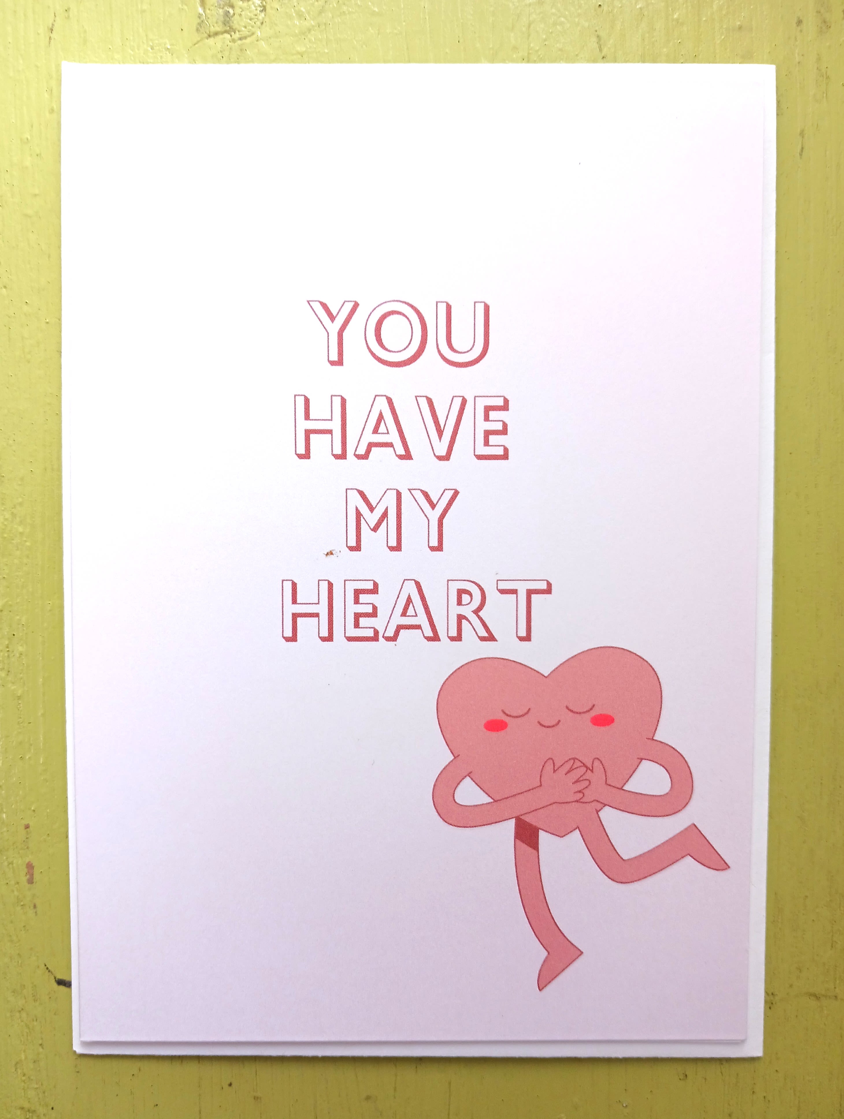 White greeting card with pink text 'You Have My Heart' and a smiling pink heart character hugging itself.