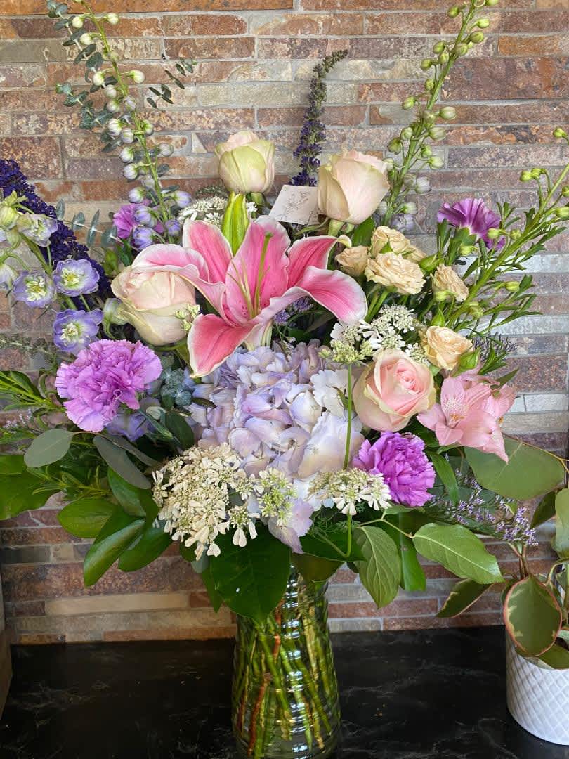 Mixed pastel bouquet with lilies, roses, hydrangea, and purple accents in a glass vase