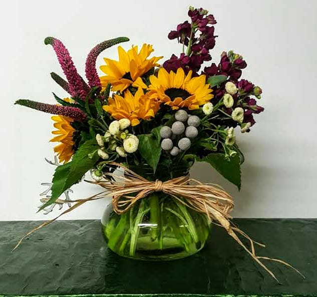 Sunny Posy - A brilliantly colored bouquet filled with spring flowers that will fill the room with wonderful fragrance and cheer.  Sunflowers, purple Stock, pink Veronica, Brunia and Chamomile (or similar flowers) fill this adorable vase accented with Raffia. Vase color will vary.