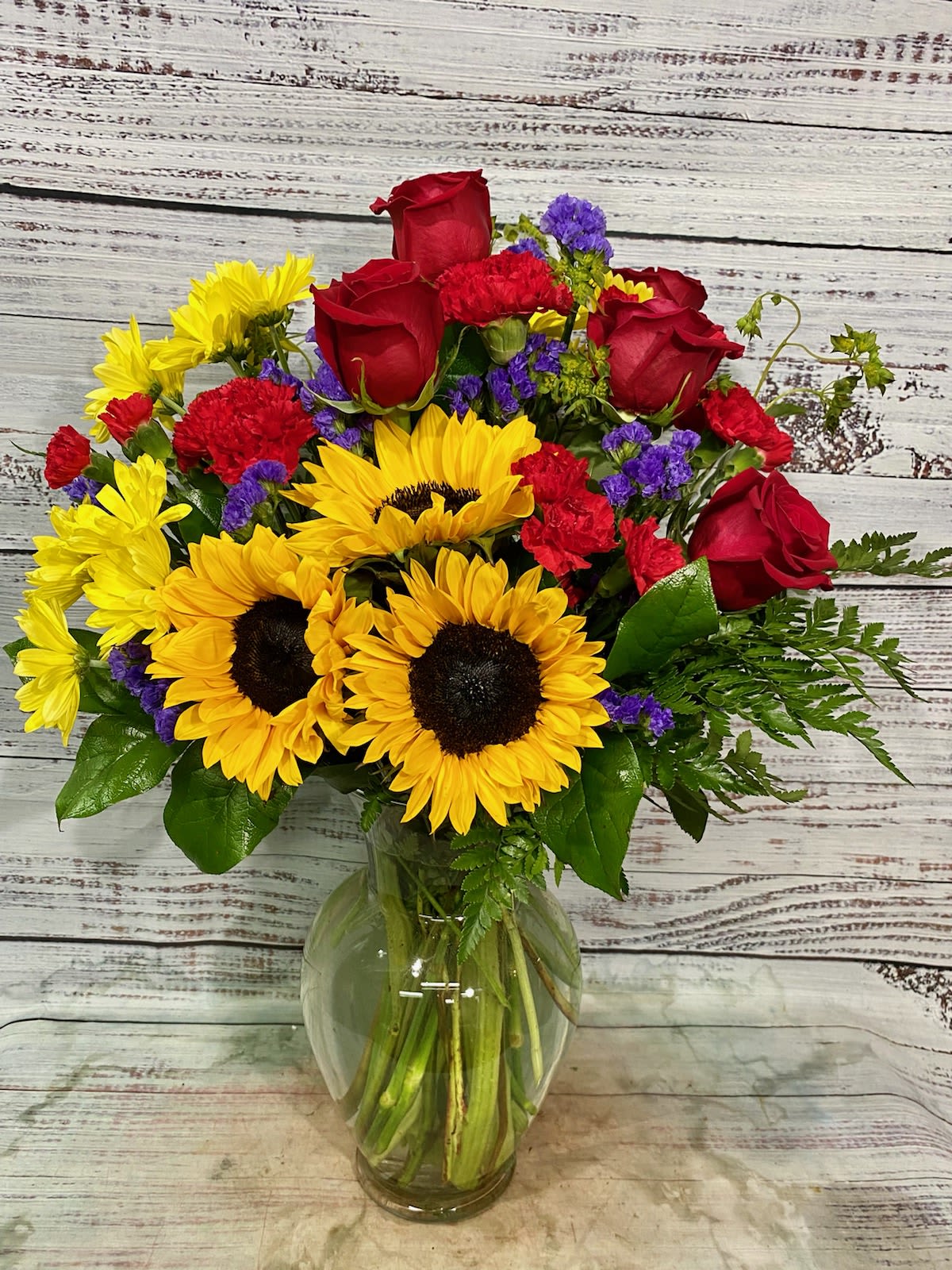 Pure Happiness - Sunny sunflowers, red roses and miniature carnations, bronze daisy spray chrysanthemums, large lavender monte cassino asters and autumn greens are beautifully arranged in a clear glass vase. Approximately 19 1/2" W x 21 1/2" H. T171-1A