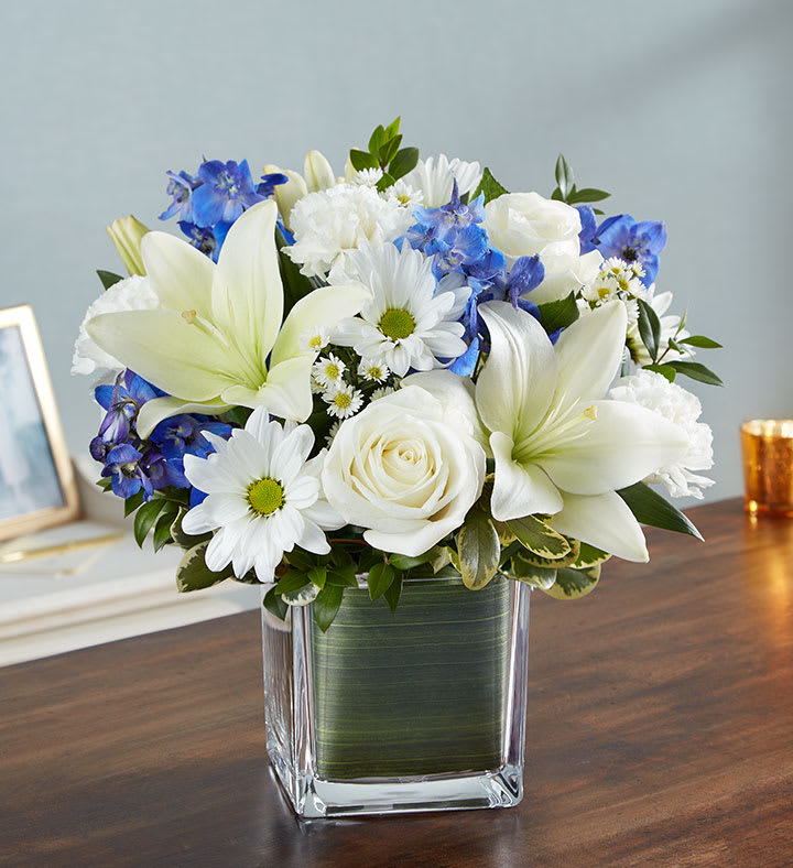 Healing Tears Blue &amp; White - Soothe their tears as they mourn the loss of a loved one with the serenity of blue and white. Our sympathy arrangement of fresh blue delphinium, white roses and lilies, expertly gathered together in a clear glass cube lined with Ti leaf ribbon, makes for an exquisite gesture of comfort and healing.
