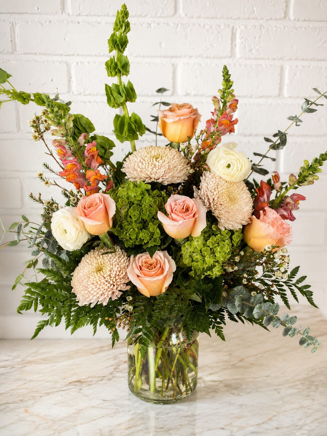 Mixed bouquet of peach roses, cream blooms, and green flowers in a glass vase