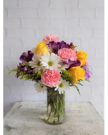 Yellow roses, pink carnations, white daisies, and purple blooms in a clear glass vase