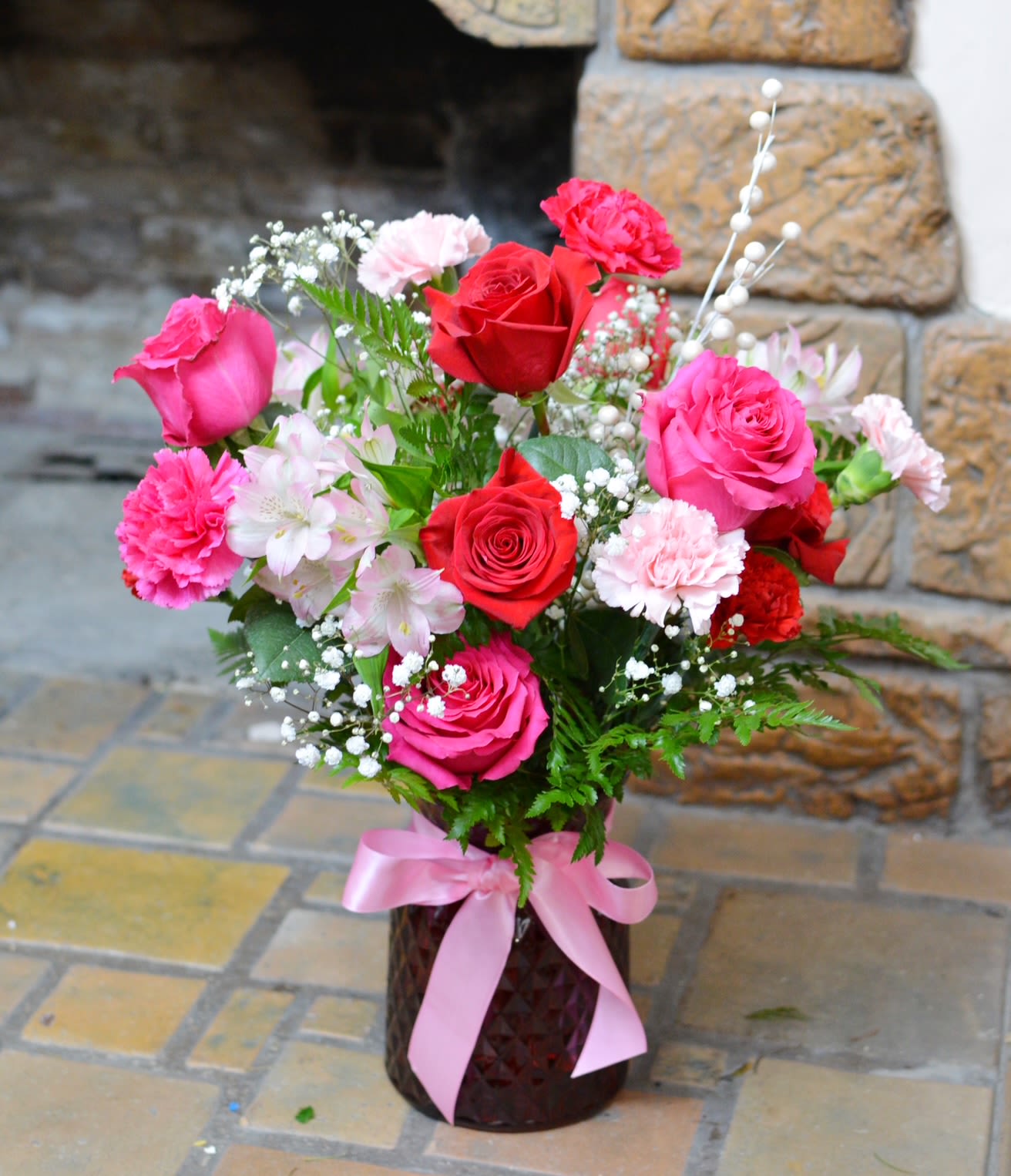 Pink and red roses with carnations in a vase tied with a pink ribbon.