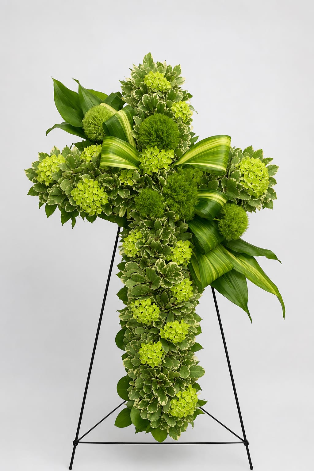 Renewal - Renewal is a contemporary floral cross that celebrates hope, remembrance, and the enduring cycle of life. Fresh green hydrangeas, textured Green Trick dianthus, variegated foliage, and sculptural tropical leaves create a design that feels peaceful, modern, and full of quiet strength. Rather than focusing on loss, Renewal speaks of love that continues, cherished memories, and new beginnings.