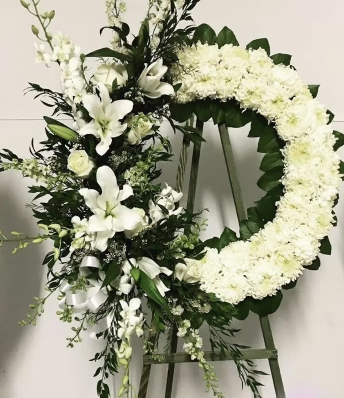 Eternal Grace Tribute - Eternal Grace Tribute is a refined and sophisticated sympathy arrangement that combines a classic circular wreath with a flowing vertical spray. Designed in soft white tones, it symbolizes peace, remembrance, and eternal love. The wreath is delicately composed of premium white chrysanthemums, representing honor and loyalty, while the cascading spray features elegant white lilies, roses, orchids, and seasonal accent blooms. Lush greenery adds depth and natural movement, creating a balanced and artistic composition. The arrangement is displayed on a handcrafted wooden easel, adding height, presence, and a polished presentation suitable for funeral services, memorials, or graveside ceremonies. This design offers a timeless and dignified tribute, expressing heartfelt sympathy with grace and elegance.