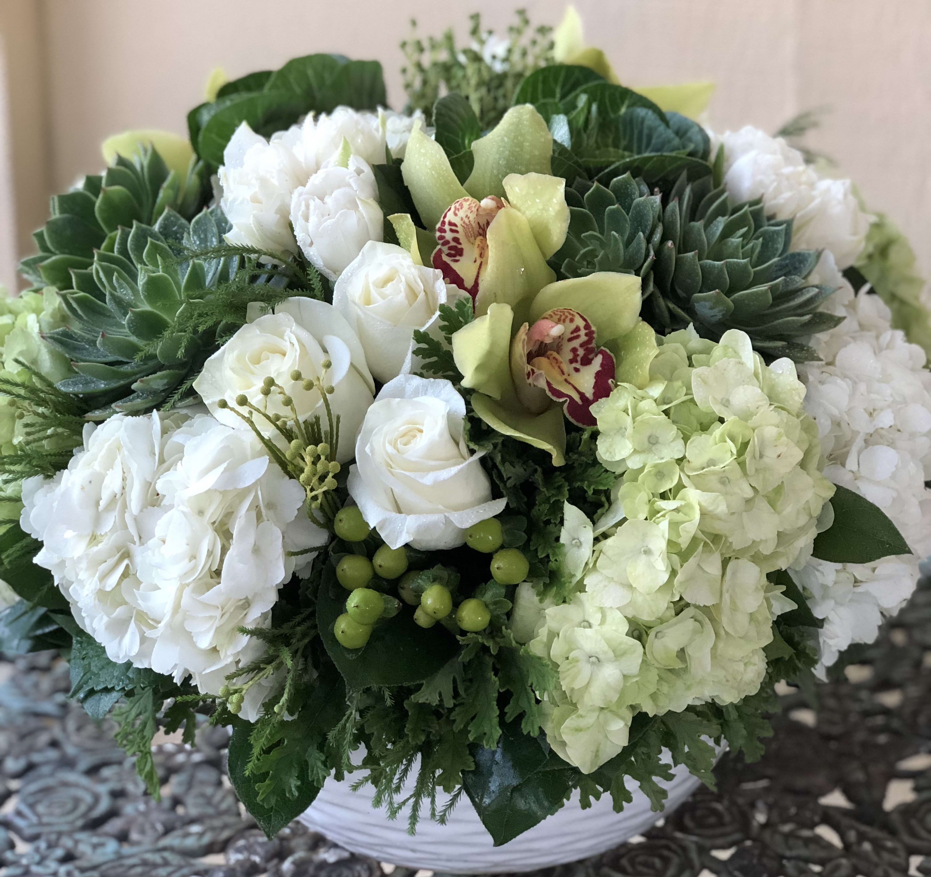Low round arrangement of white roses, hydrangeas, green succulents, and orchids in a white bowl