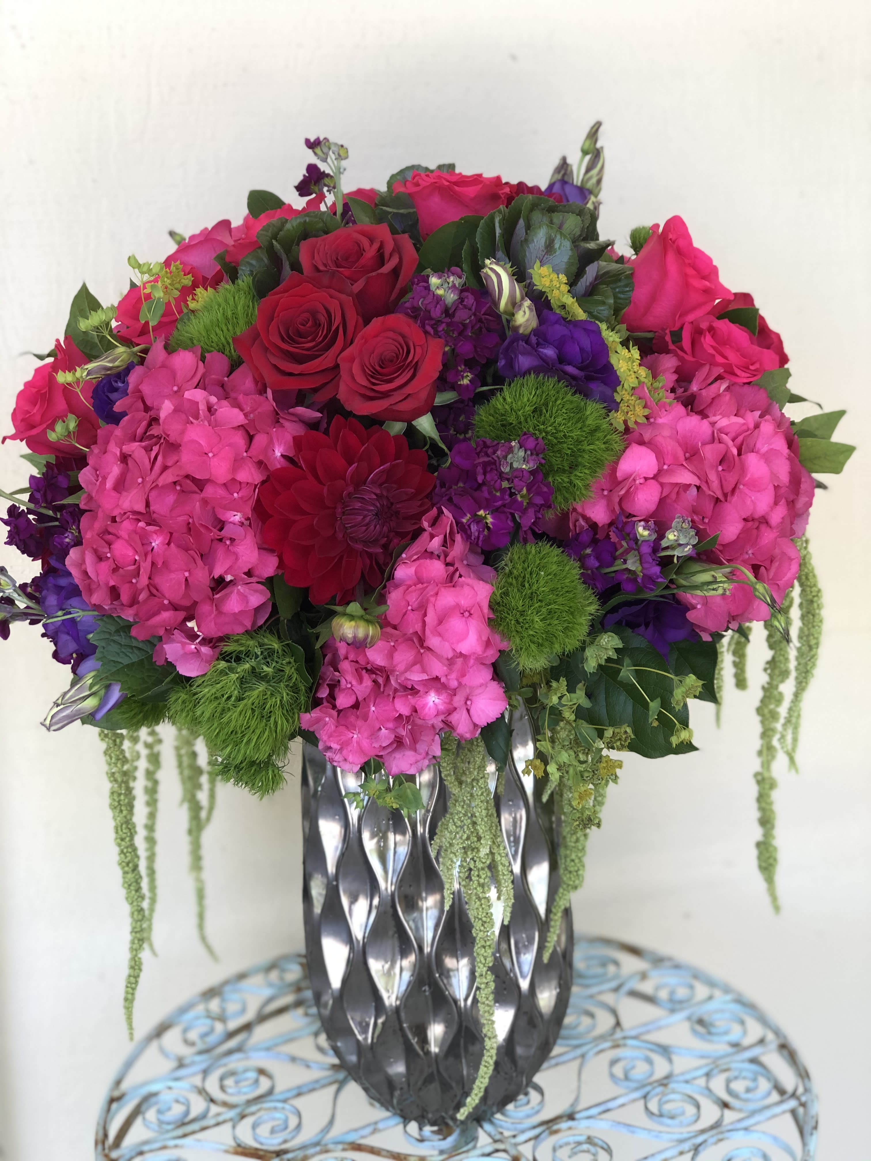 Tall arrangement of hot pink hydrangeas, red roses, and purple blooms in a silver patterned vase