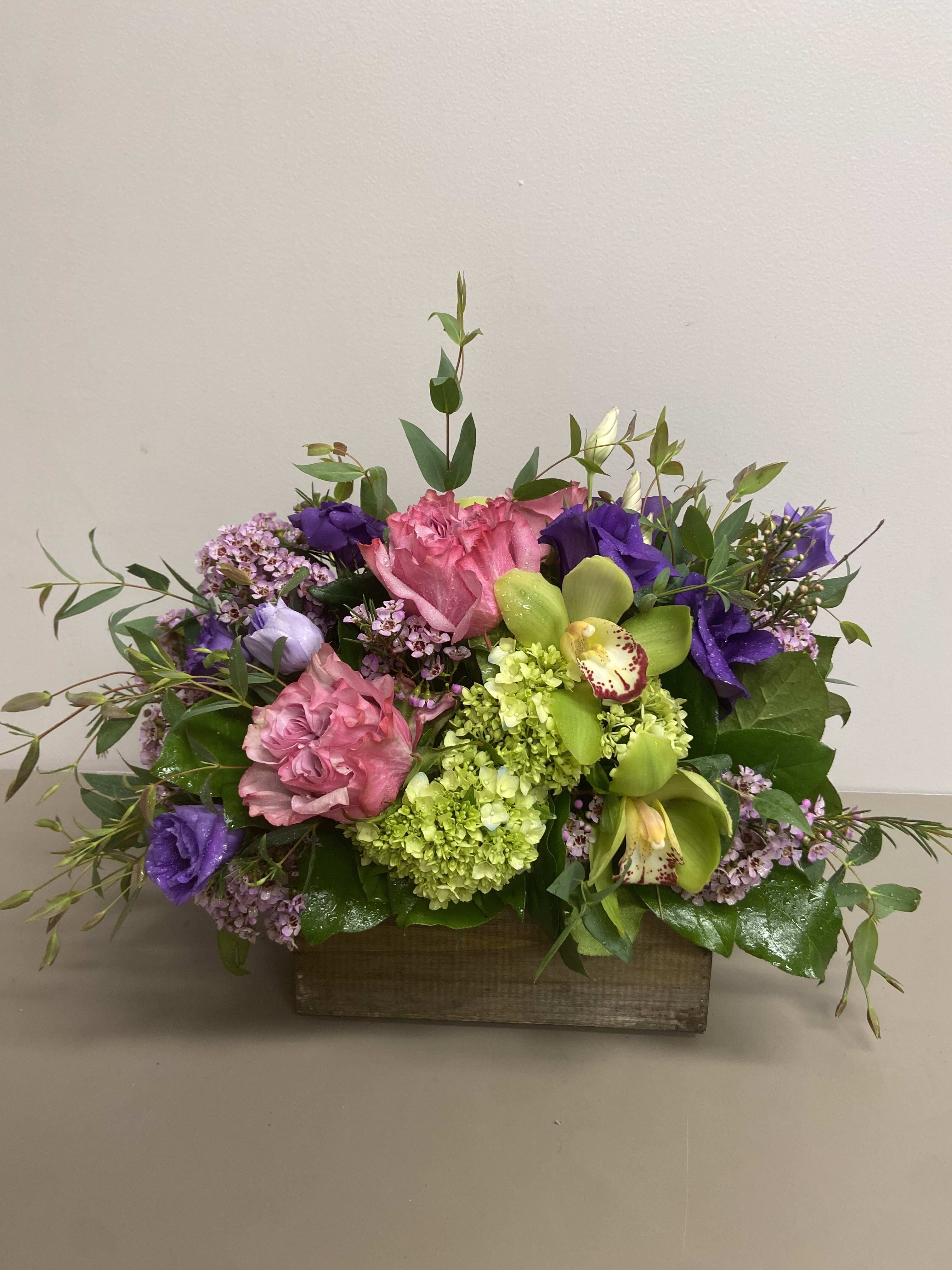 Low arrangement of pink roses, green hydrangeas, orchids and purple blooms in a rectangular wooden box