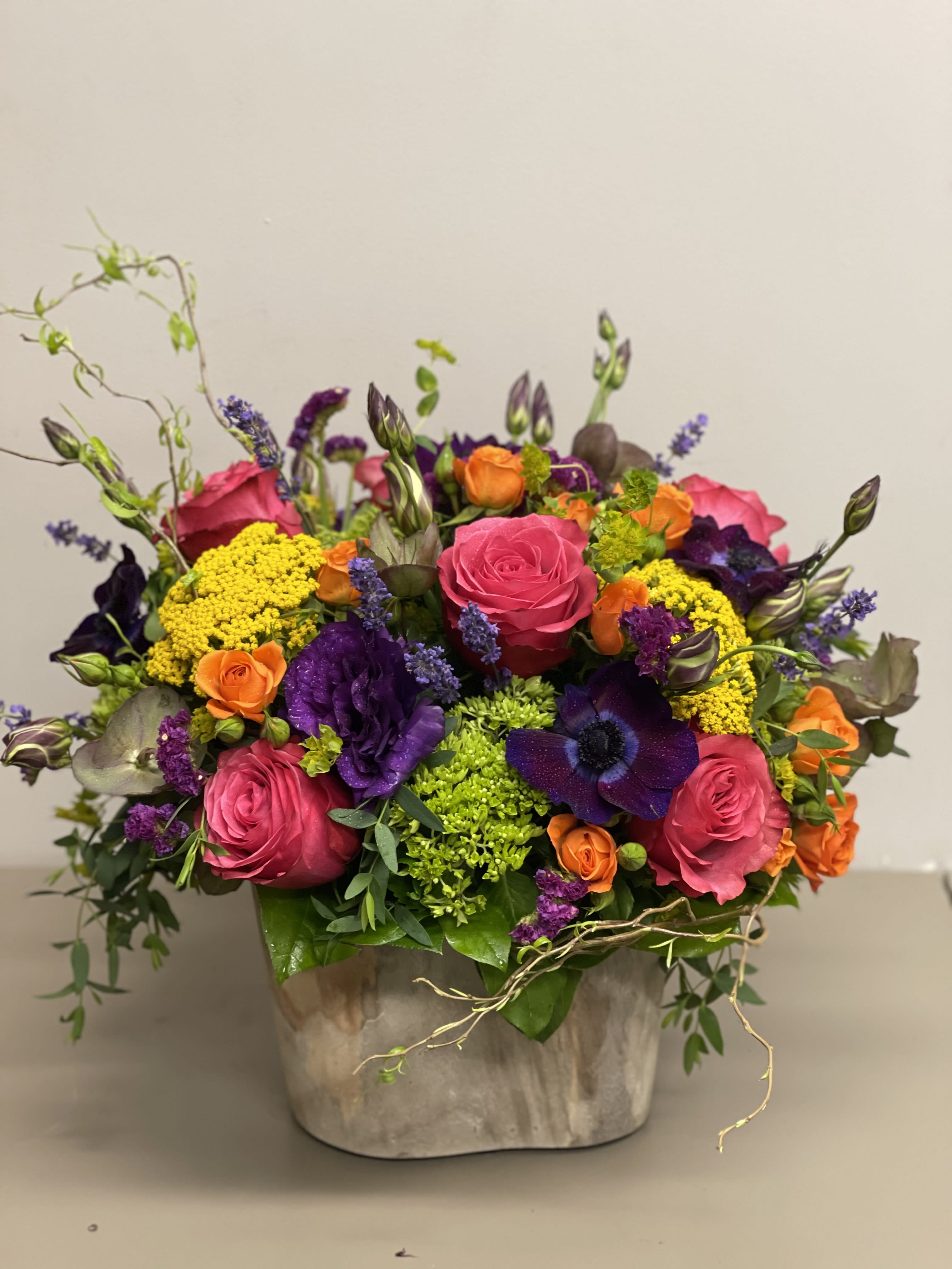 Vibrant arrangement of pink roses, purple blooms, orange flowers and green accents in a rustic ceramic container.