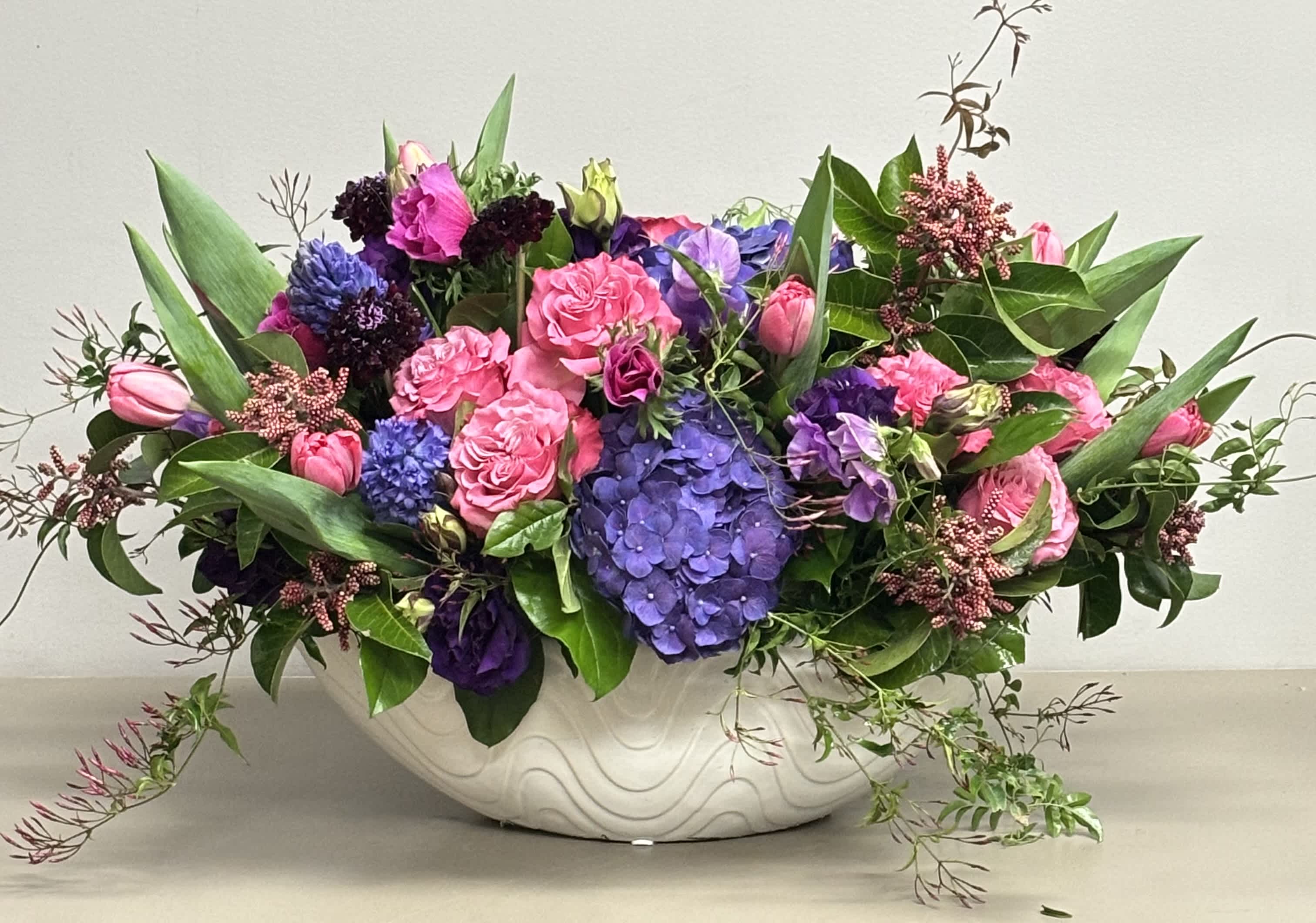 Low oval arrangement of pink roses and tulips with purple hydrangeas in a white ceramic bowl