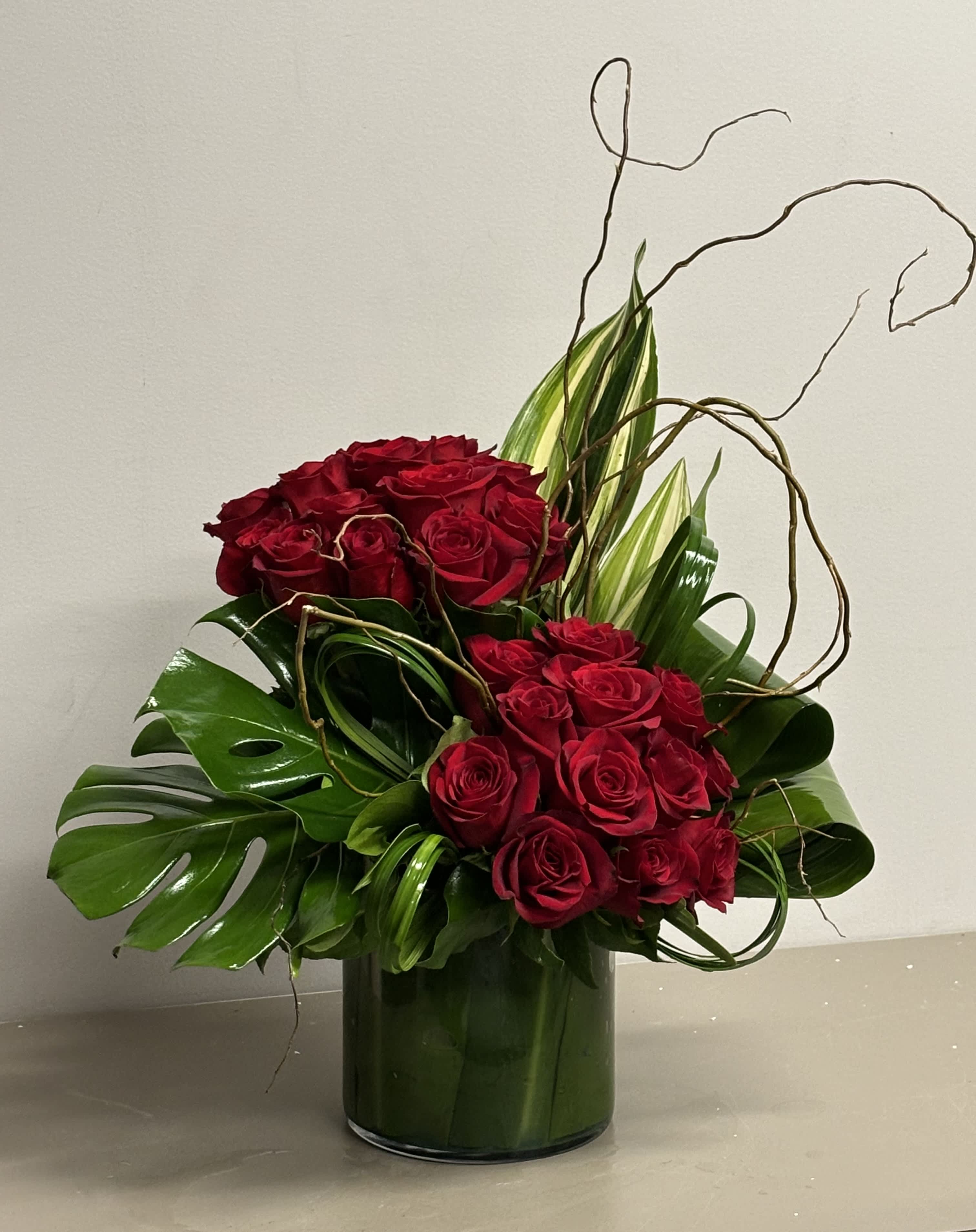 Tall arrangement of red roses with tropical green leaves and curly branches in a clear glass cylinder vase