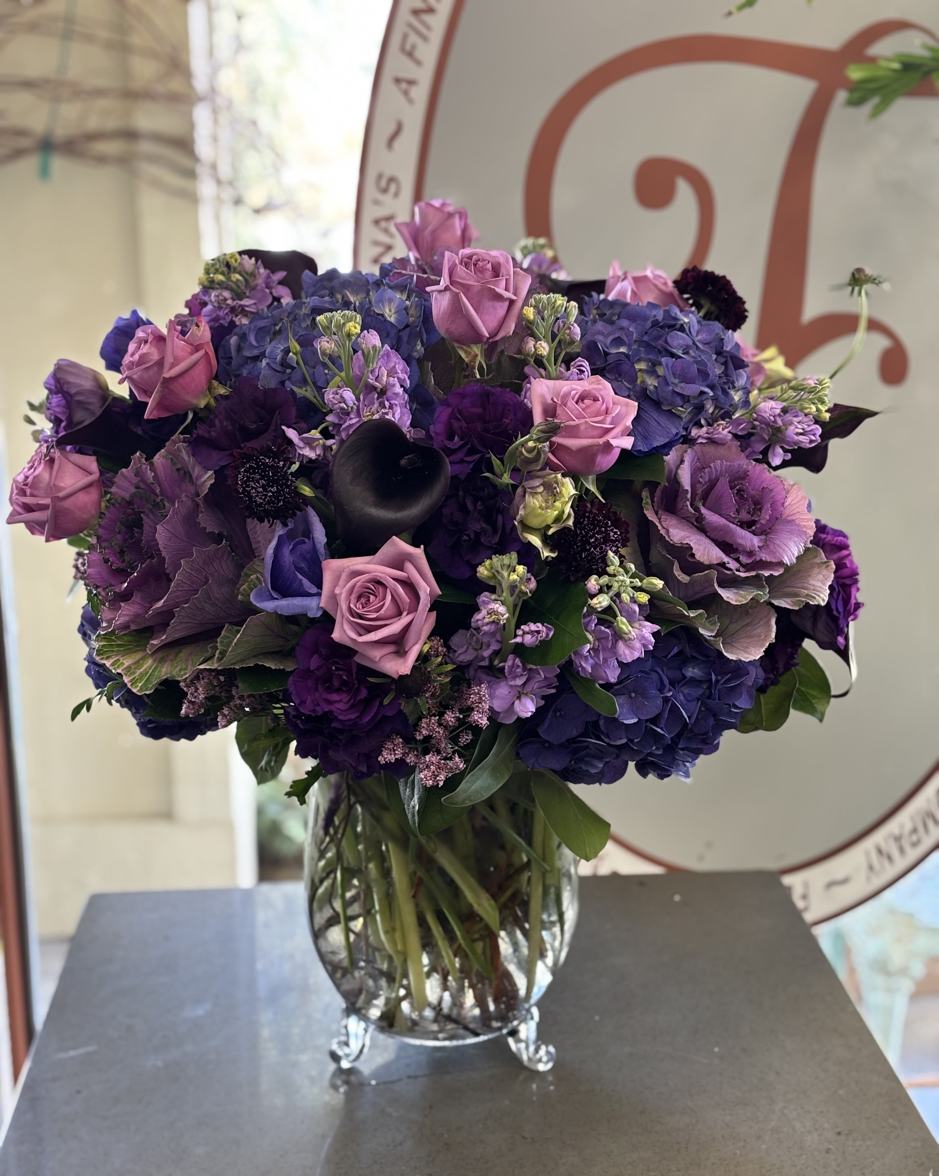 Lavender roses and purple hydrangeas arranged in a clear footed glass vase