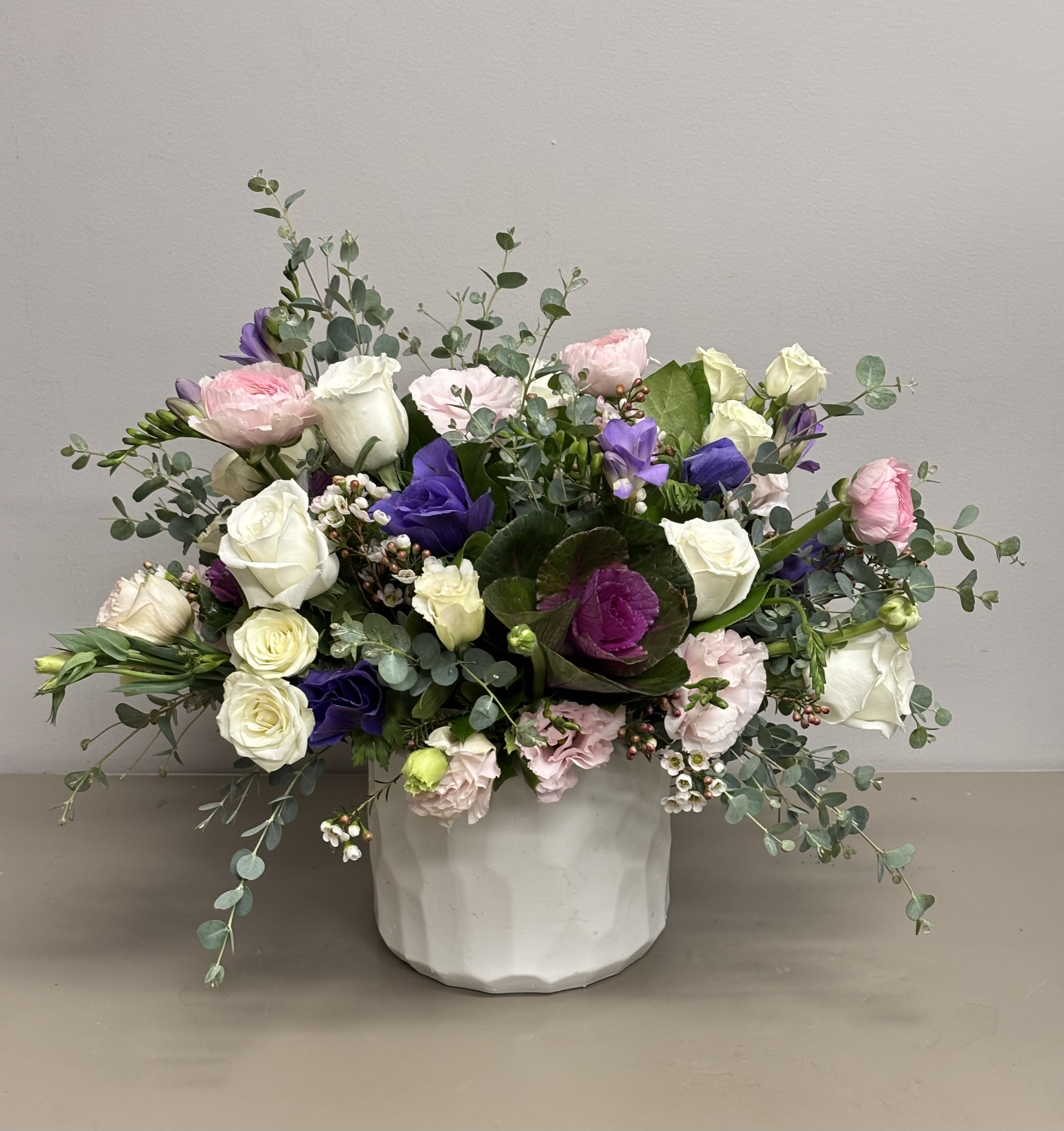 Low arrangement of white roses, pink blooms, and purple flowers in a white ceramic pot