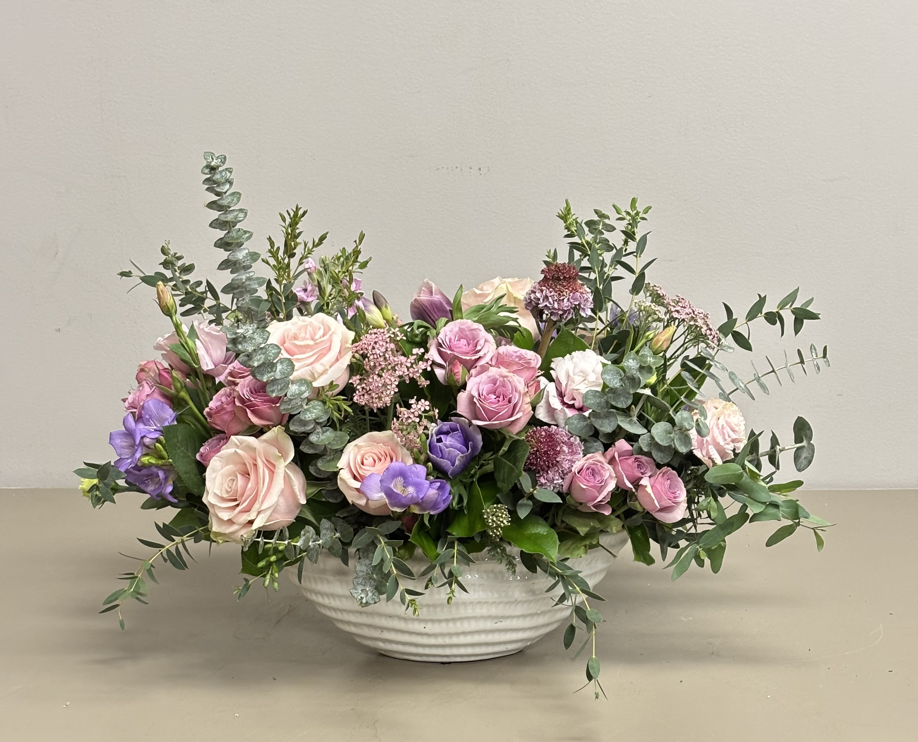 Low centerpiece of pink roses and mixed purple flowers in a white ceramic bowl