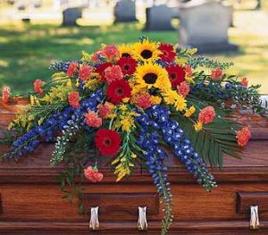 Bright mixed casket spray with sunflowers, red gerbera daisies, and blue flowers on a wooden casket