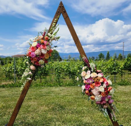Triangle wooden arch decorated with pink, peach, and white flower clusters in an outdoor field
