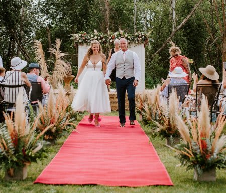 Outdoor wedding aisle with pampas grass arrangements and a flower-topped arch behind a couple on a red carpet.