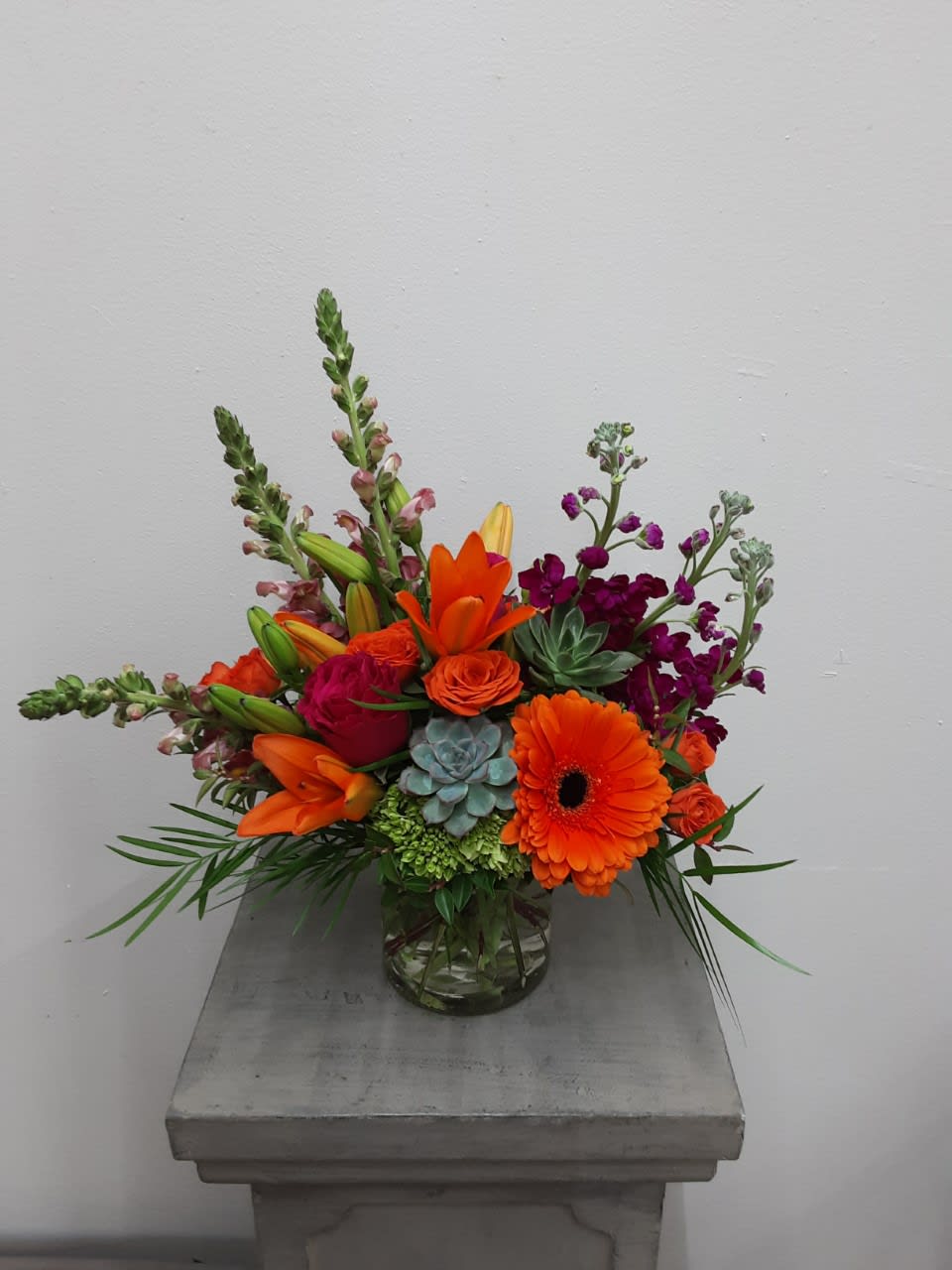 Bright mixed arrangement with orange gerbera, lilies, roses, and succulents in a clear glass vase