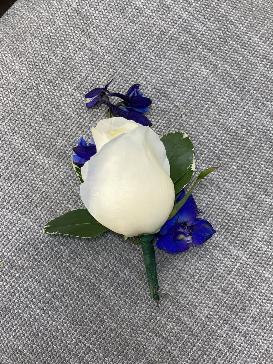 White rose boutonniere with blue accent flowers on gray fabric