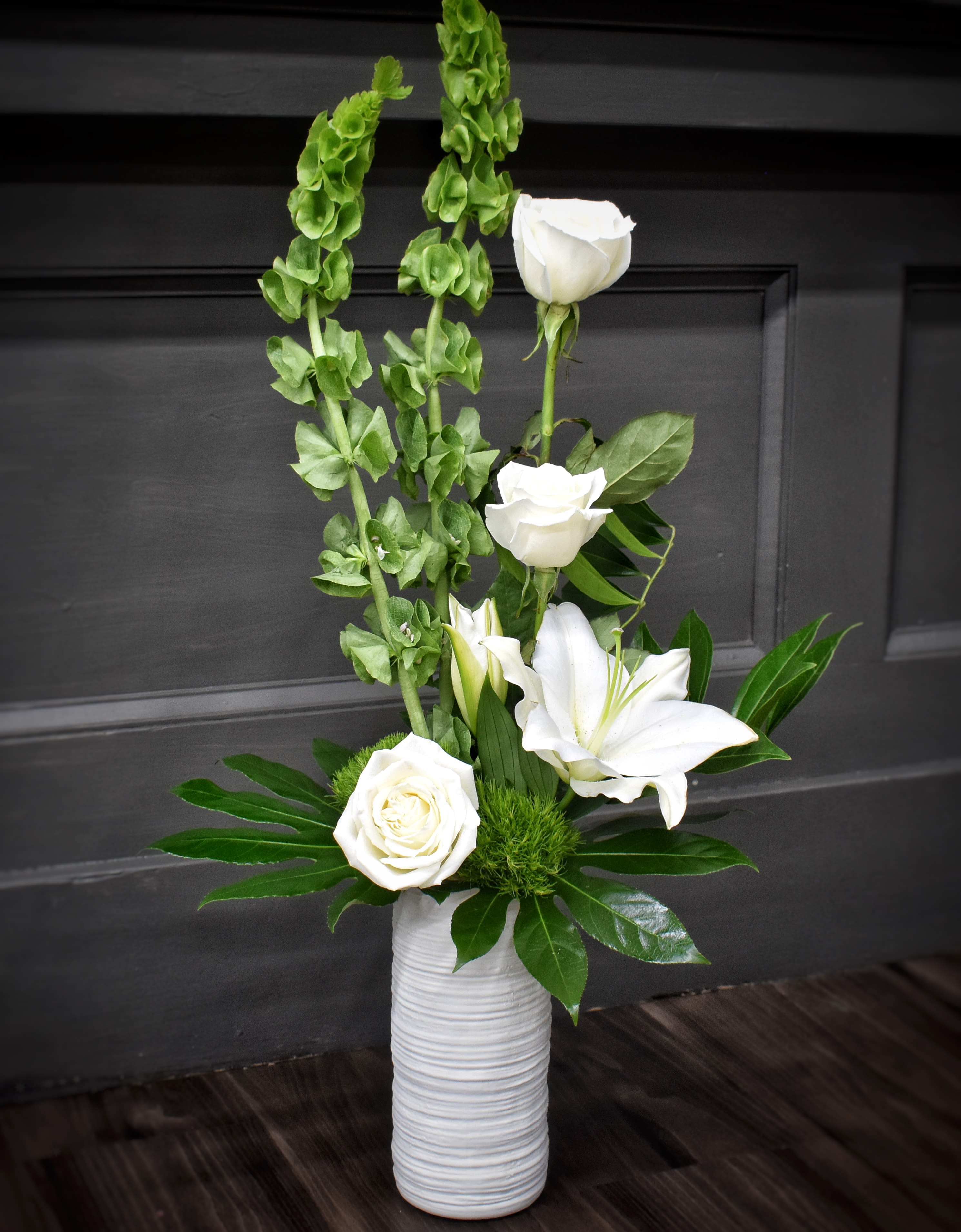 Peaceful Wishes - Pure white lilies and roses rise from tall green spikes in a vertical, airy arrangement-an elegant flower arrangement for sympathy, a wedding, or any moment that calls for calm, refined beauty. The design's clean white-and-green palette and formal silhouette make the recipient feel held and honored, the kind of gift that reads as both peaceful and purposeful.  Perfect for offering comfort, marking a wedding, or sending thoughtful congratulations, this arrangement arrives ready to display in its white ceramic vessel. Its vertical profile makes a serene statement in smaller spaces-no special care beyond fresh water is needed to keep the blooms looking pure and radiant.