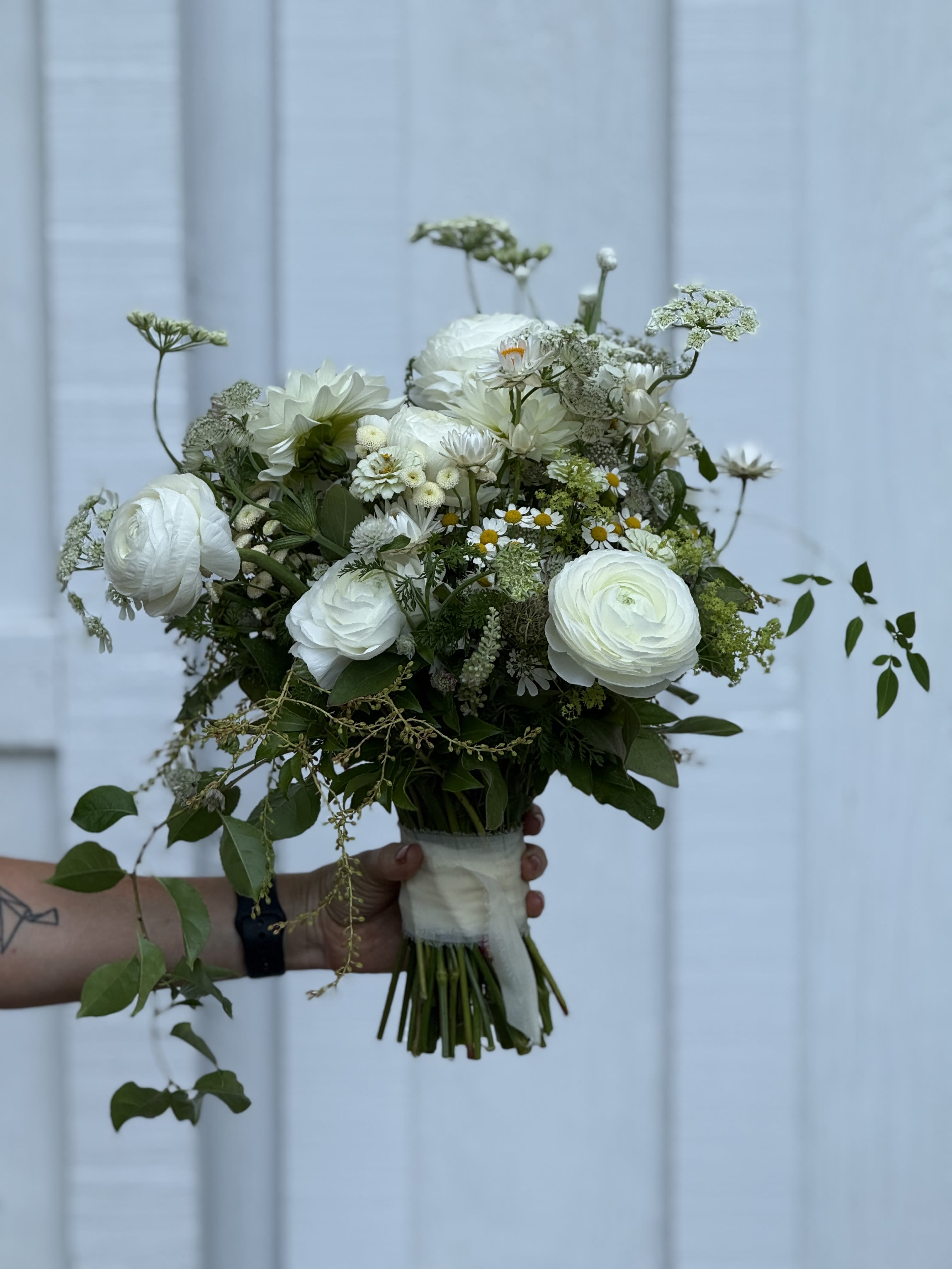 Ala carte Bridal bouquet  - A soft, garden-fresh bouquet that reads effortlessly bridal bouquet is designed for weddings and elopements when you want a romantic, organic look. The mostly-white palette with touches of green and mixed-color accents creates a luminous, classic feel that suits ceremony or reception settings; it's the kind of bouquet that makes the person you love look like they just stepped out of a sunlit garden.  The arrangement is loose and hand-tied, combining large ranunculus-style blooms with small daisy-style flowers, delicate filler blossoms, and trailing greenery for airy texture and movement. Stems are wrapped with a pale ribbon for an understated finish that complements white, cream, and soft-colored gowns. The mixed-bouquet construction keeps the silhouette natural rather than overly structured, giving gentle contrast between rounded blooms and wispy foliage.  Perfect for brides seeking a romantic, organic statement or for attendants who want coordinating bouquets with a soft color story, this piece is easy to carry and photographs beautifully from every angle. Specify ribbon color and any color preferences at checkout to customize the look.