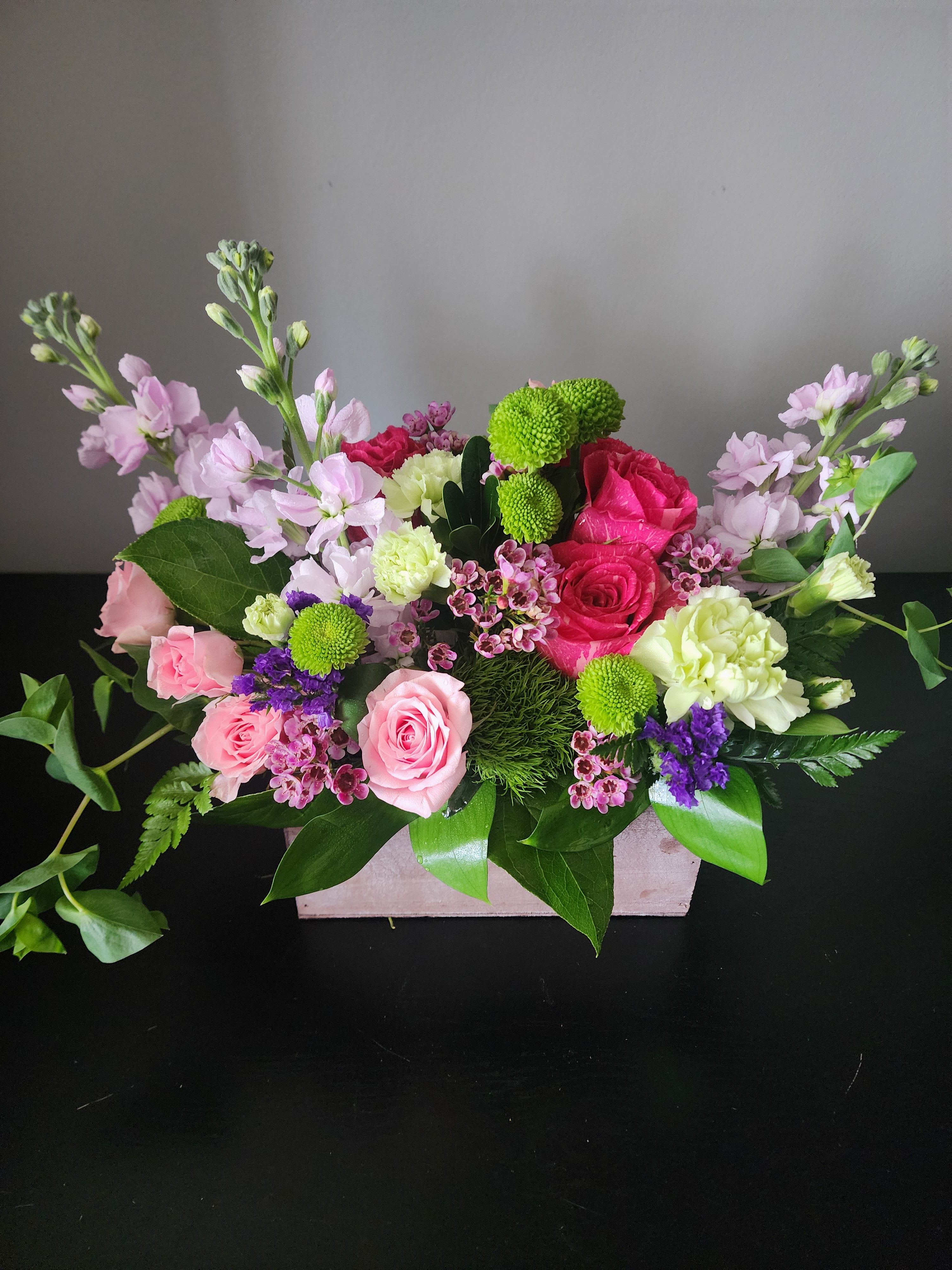 Blushwood Blooms by MG Florist  - Blushwood Blooms | The Magic Garden Florist  Elevate any space with our rectangular wooden box floral arrangement. exclusively crafted by The Magic Garden Florist, Rochester, NY. This elegant design showcases a harmonious blend of fresh pink roses, spray roses, green button mums, soft carnations, and fragrant lavender stock, beautifully complemented by delicate waxflower, statice, and lush seasonal greenery.  Accented with unique textures like trick dianthus, this arrangement offers a stunning balance of color, depth, and natural charm—making it perfect for birthdays, anniversaries, get well gifts, or home décor.  Handcrafted with care, this premium floral arrangement is designed to impress with its **long-lasting freshness**, **modern rustic presentation**, and **garden-inspired elegance**.  * Premium fresh-cut pink roses and seasonal blooms * Stylish rectangular wooden box container * Soft pastel and vibrant green color palette * Perfect for gifting, events, or interior decoration * Hand-arranged by expert florists  Whether you're searching for a pink rose arrangement, or a unique floral gift, this design brings timeless beauty and sophistication to any occasion.  Order now from The Magic Garden Florist for same day delivery and experience the art of floral design delivered with elegance. 