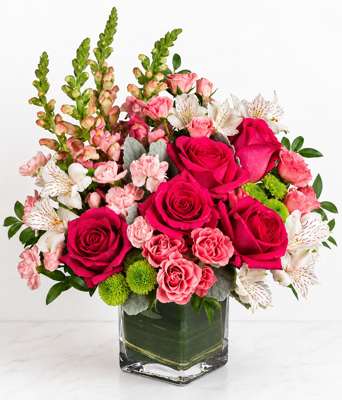 Joyful Bouquet - Hot-pink roses clustered with soft pink carnations and crisp white alstroemeria create an instantly joyful birthday bouquet or anniversary arrangement that will make them feel cherished. This pink, white and green flower arrangement brings a romantic yet celebratory mood-perfect for birthdays, Mother's Day, anniversaries, or simply saying "I'm thinking of you" to someone special.  Tall green snapdragon spikes add elegant height and structure behind the lush cluster of blooms, while small green chrysanthemum pompons and silvery foliage give fresh contrast to the saturated pinks. Arranged in a clear square glass vase, the composition reads full and layered from every angle; the mix of textures-from silky rose petals to the feathery snapdragons-creates a vibrant, tactile display.  This is the kind of bouquet that helps the recipient feel genuinely celebrated and seen. Available in a compact square vase that fits tabletops and mantels, it arrives ready to display; change the water every few days and trim stems slightly to extend the life of the flowers.