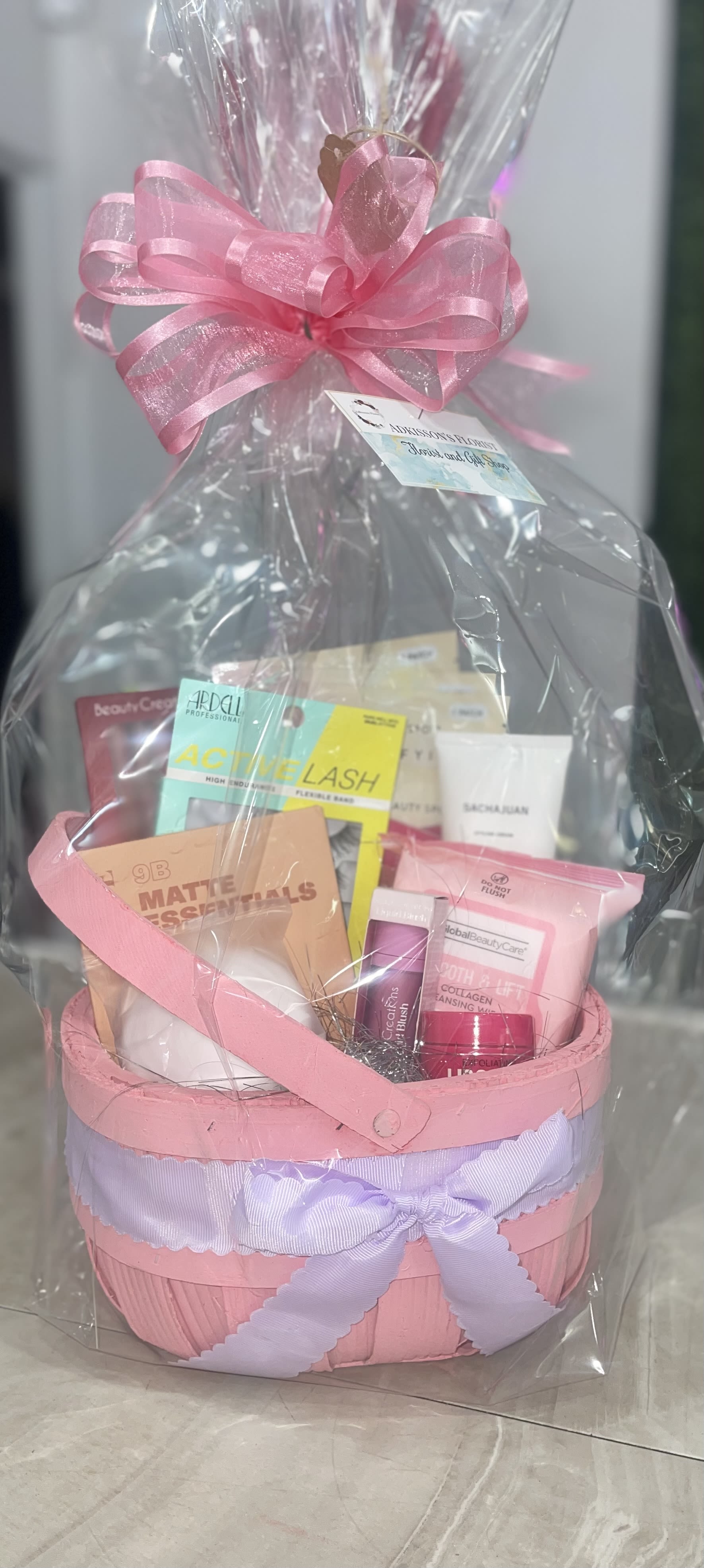 Makeup Lover Gift Basket  - Soft pastels and pampering treats make this Makeup Lover Gift Basket an unforgettable birthday or thank-you gift that reads like a personal spa day. The gift set is presented as a non-floral arrangement ideal for an anniversary, birthday, or any occasion when you want the person you're thinking of to feel truly spoiled-she'll open a bundle of beauty picks and feel seen.  Inside a pink or light-blue woven basket the assortment includes boxed and tube-style cosmetics, skincare jars and packets, a makeup palette, cosmetic tubes, false eyelashes, a small jar, hair accessory, folded towel, and other curated beauty product packages. The basket is wrapped in clear cellophane and finished with a large organza or pink/lavender ribbon bow, with sizes and container style varying by colorway so each presentation stays fresh and gift-ready.  Perfect for someone who loves makeup, skincare, or a little everyday luxury, this ready-to-give set works as a birthday surprise, a thoughtful thank-you, or a lovely anniversary treat. No assembly required - simply select your preferred colorway at checkout and we'll deliver a neatly wrapped basket that's ready to place in their hands.