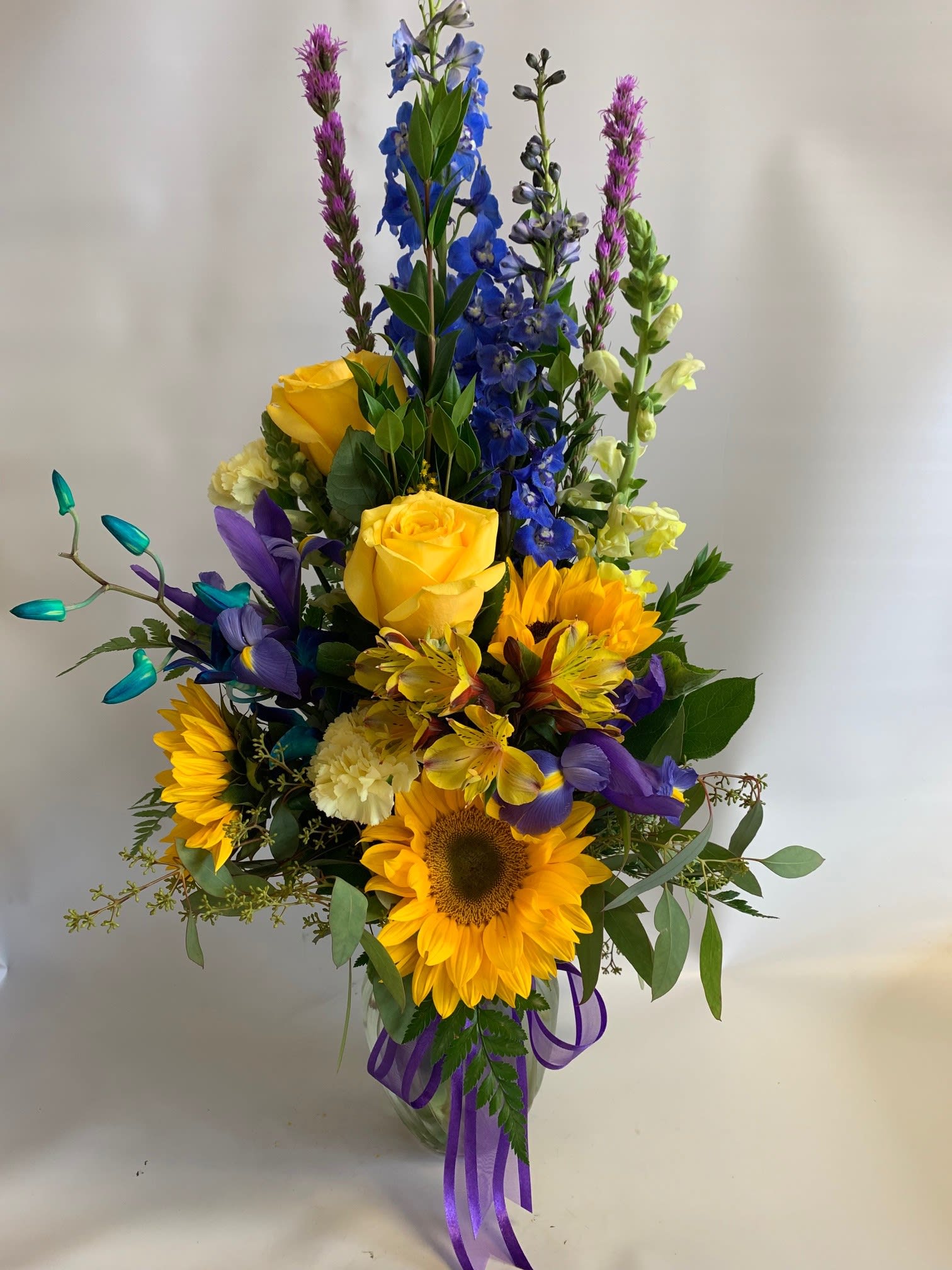 Tall mixed bouquet with sunflowers, yellow roses, and blue flowers in a glass vase