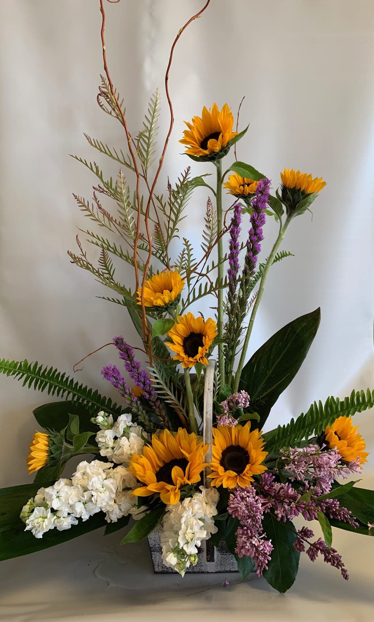 Tall floral arrangement with sunflowers, white blooms, and purple flowers in a square container