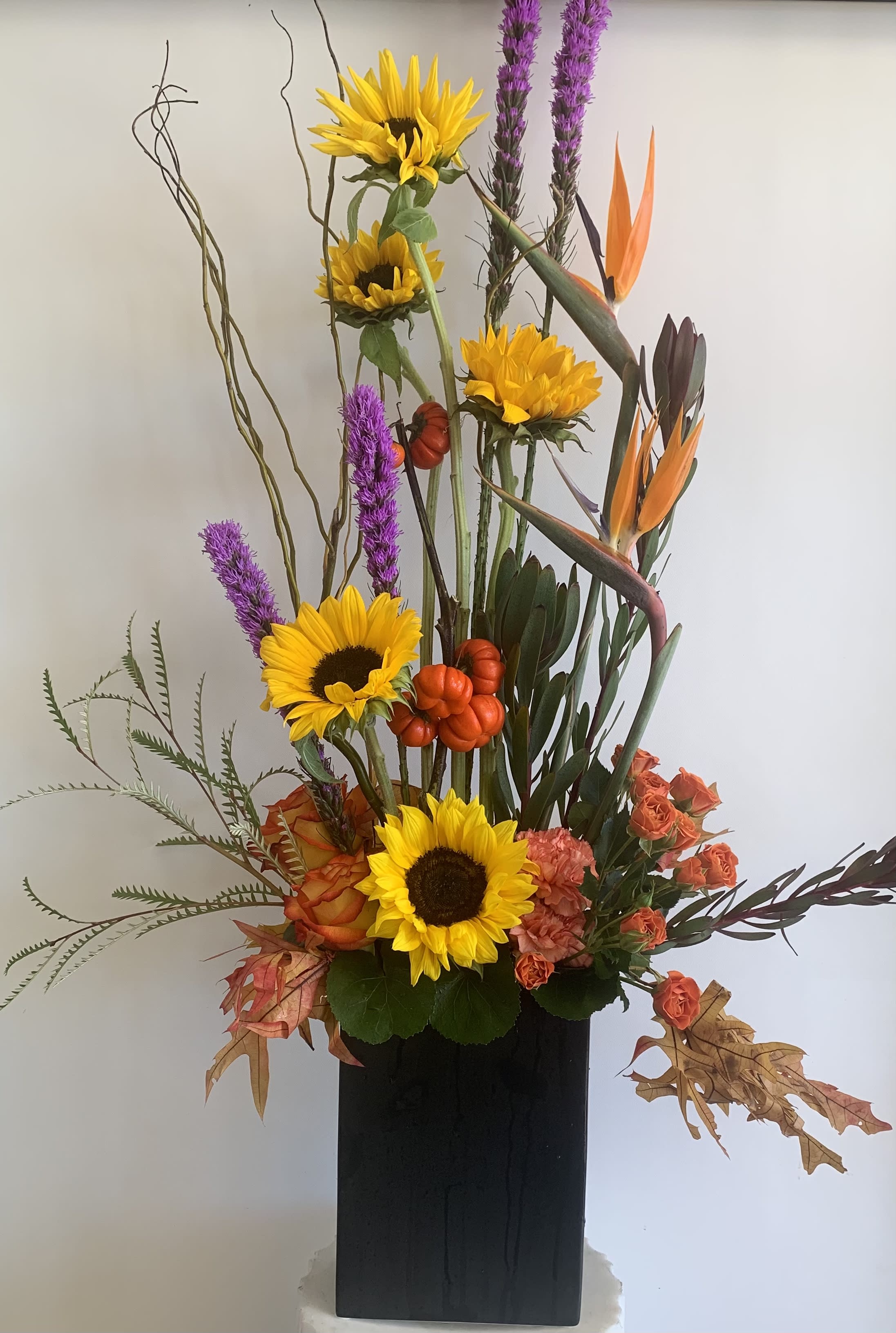 Tall floral arrangement with sunflowers, orange blooms, and purple spikes in a black vase