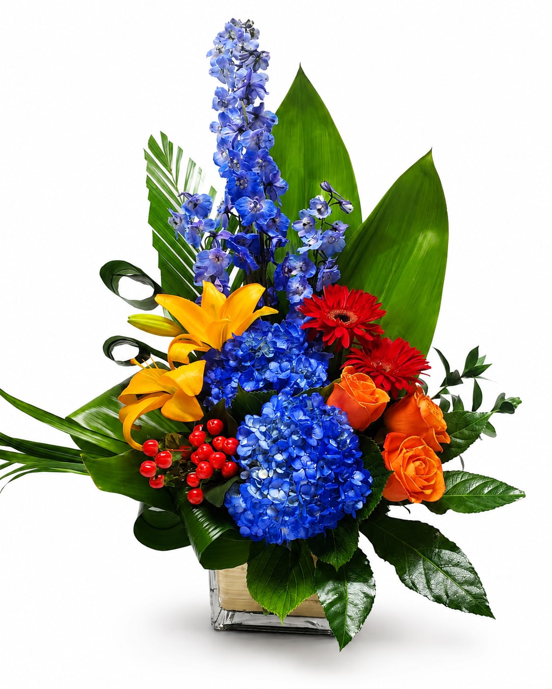 Beauty &amp; Bold - A bold and colorful floral design featuring brilliant blue hydrangeas and delphinium, vibrant orange lilies and roses, striking red gerbera daisies, and festive red berries, accented with lush tropical greenery. Arranged in a modern glass cube vase, this eye-catching centerpiece adds energy, elegance, and sophistication to any celebration or space.