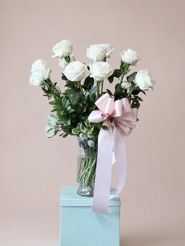 White Roses Bouquet  - White long-stemmed roses arranged in a clear glass vase create a quietly elegant birthday bouquet or anniversary roses gift that reads as both romantic and refined. The white roses' clean petals and dark green foliage give a fresh, classic look while the pale pink satin ribbon tied around the vase adds a soft, romantic contrast-perfect for saying "I love you," "happy birthday," or "congratulations" without overstating the moment.  The bouquet sits presented on a light blue gift box, making it ready for immediate gifting. Each rose is shown with long stems and generous leaves for a natural, graceful silhouette; the clear glass vase highlights stem length and keeps the focus on the blooms. Color accents of pink, green, and blue in the ribbon, foliage, and gift box make the arrangement feel thoughtful and composed.  This is the kind of bouquet that will make the recipient feel truly seen-gentle, celebratory, and understatedly luxurious. Standard care: keep vase filled with fresh water, trim stems slightly every few days, and place away from direct heat or sunlight to extend bloom life.
