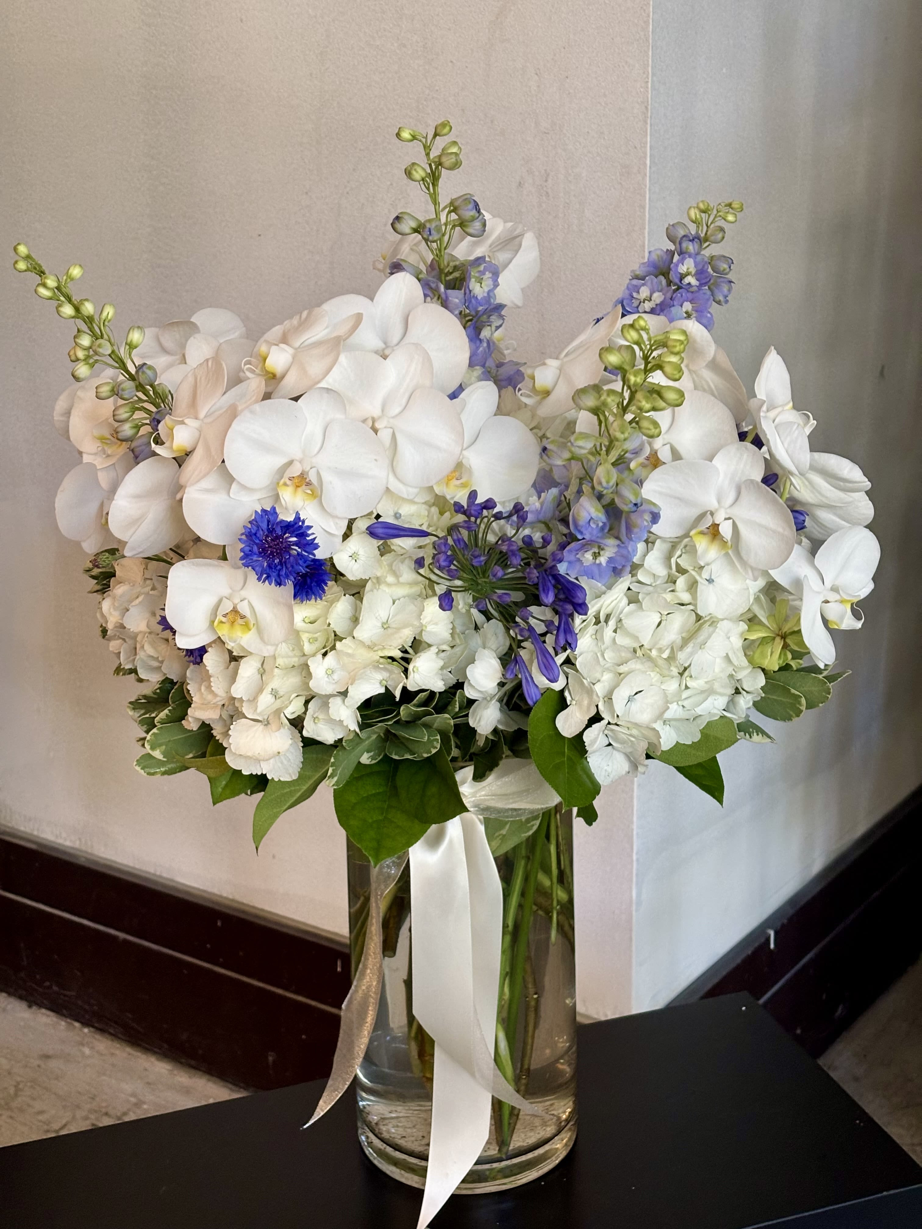 White orchids and blue flowers arranged in a tall glass vase with ribbon