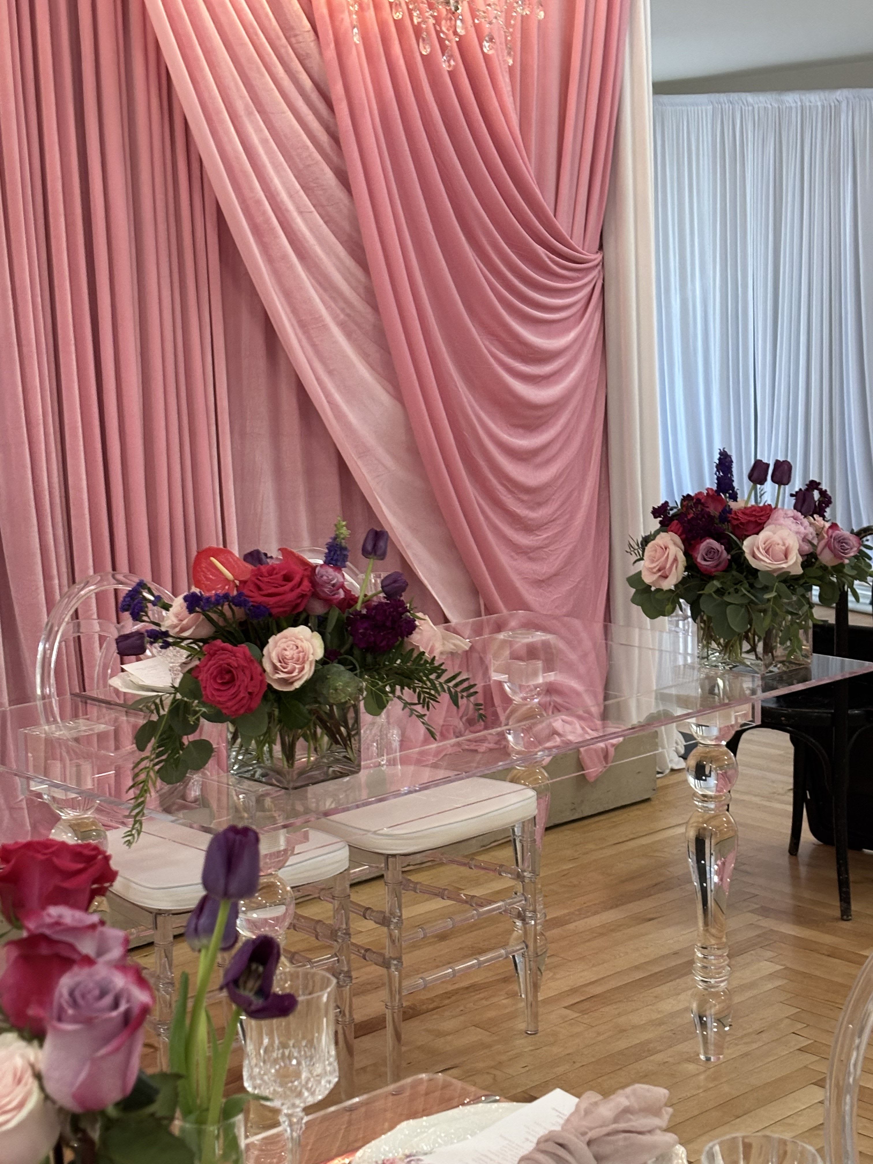 Pink and purple floral centerpieces on clear acrylic tables with draped pink fabric backdrop