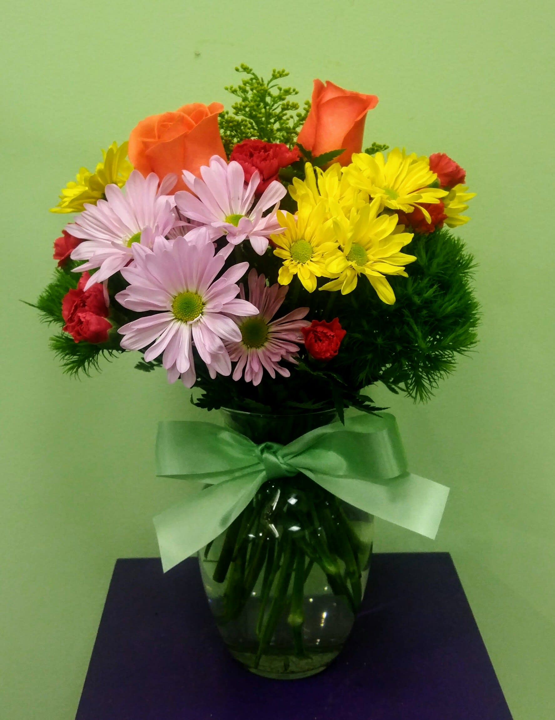 Bouquet of orange roses, daisies, and red carnations in a glass vase with a ribbon
