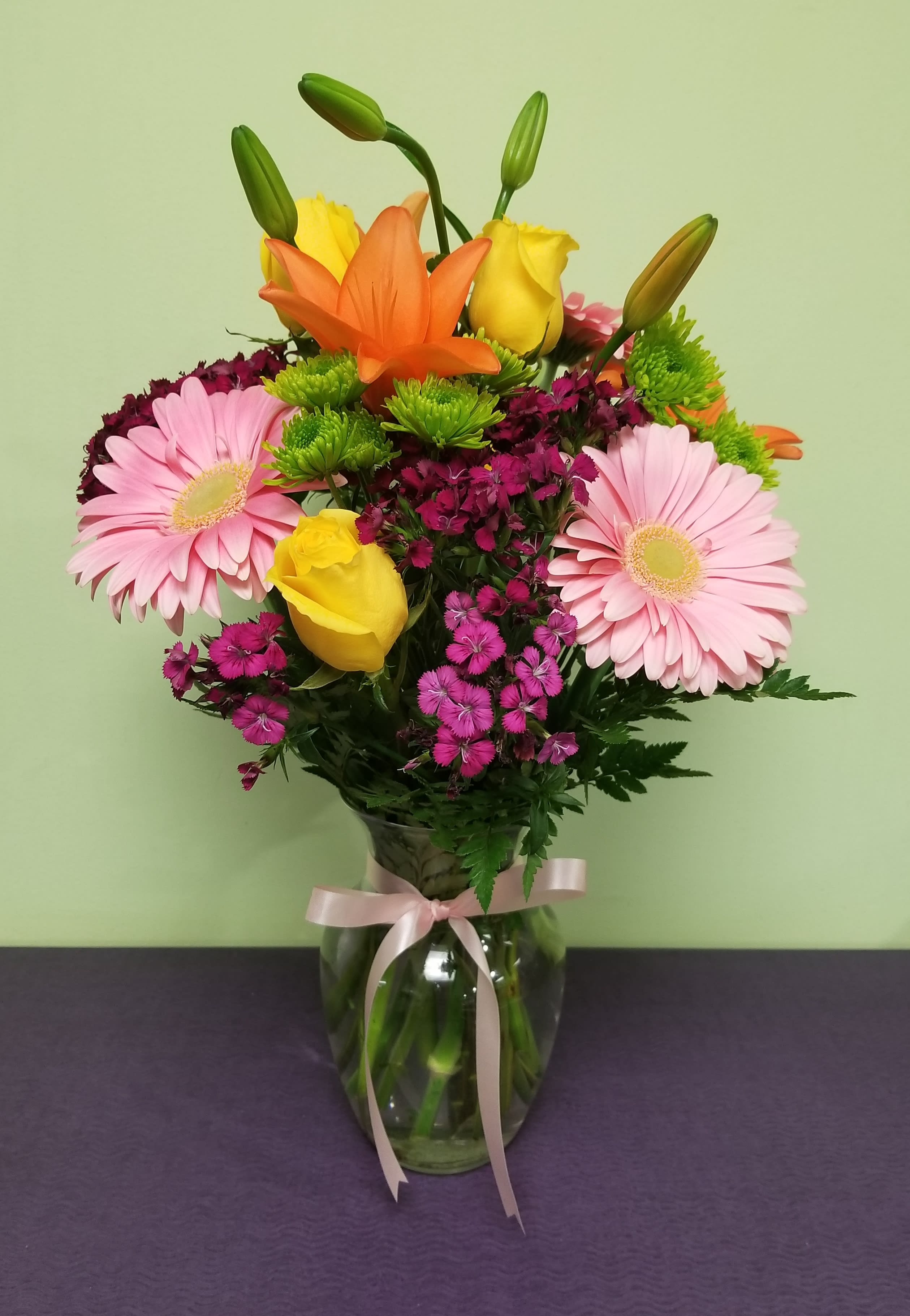 Bouquet of pink gerbera daisies, yellow roses, and orange lilies in a glass vase