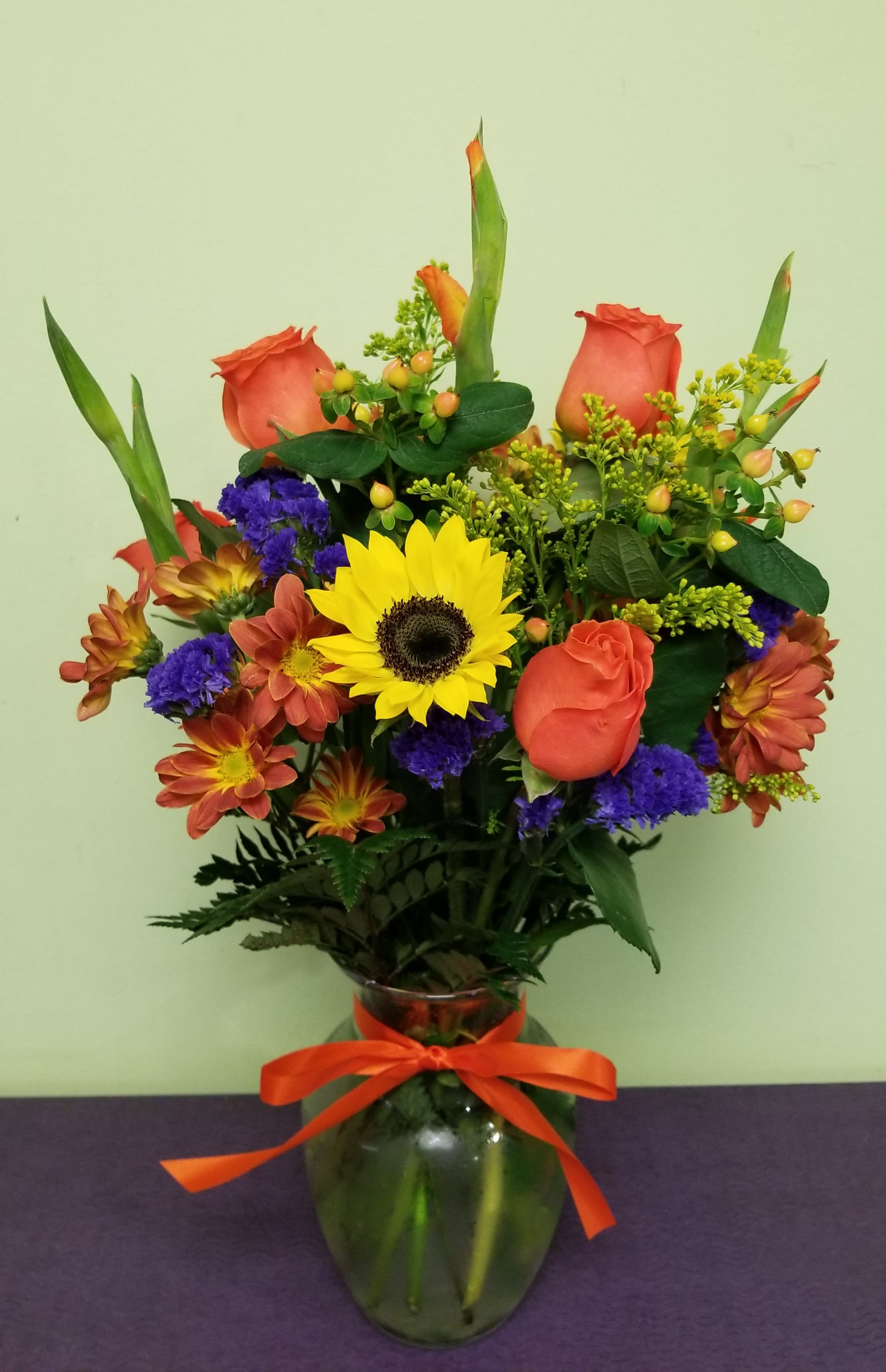 Mixed bouquet of orange roses, sunflowers, and purple flowers in a glass vase