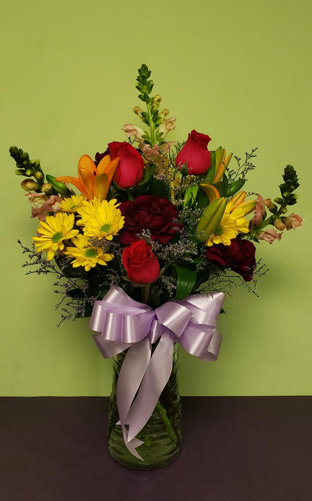 Mixed bouquet of roses, lilies, and yellow daisies in a glass vase with a lavender bow