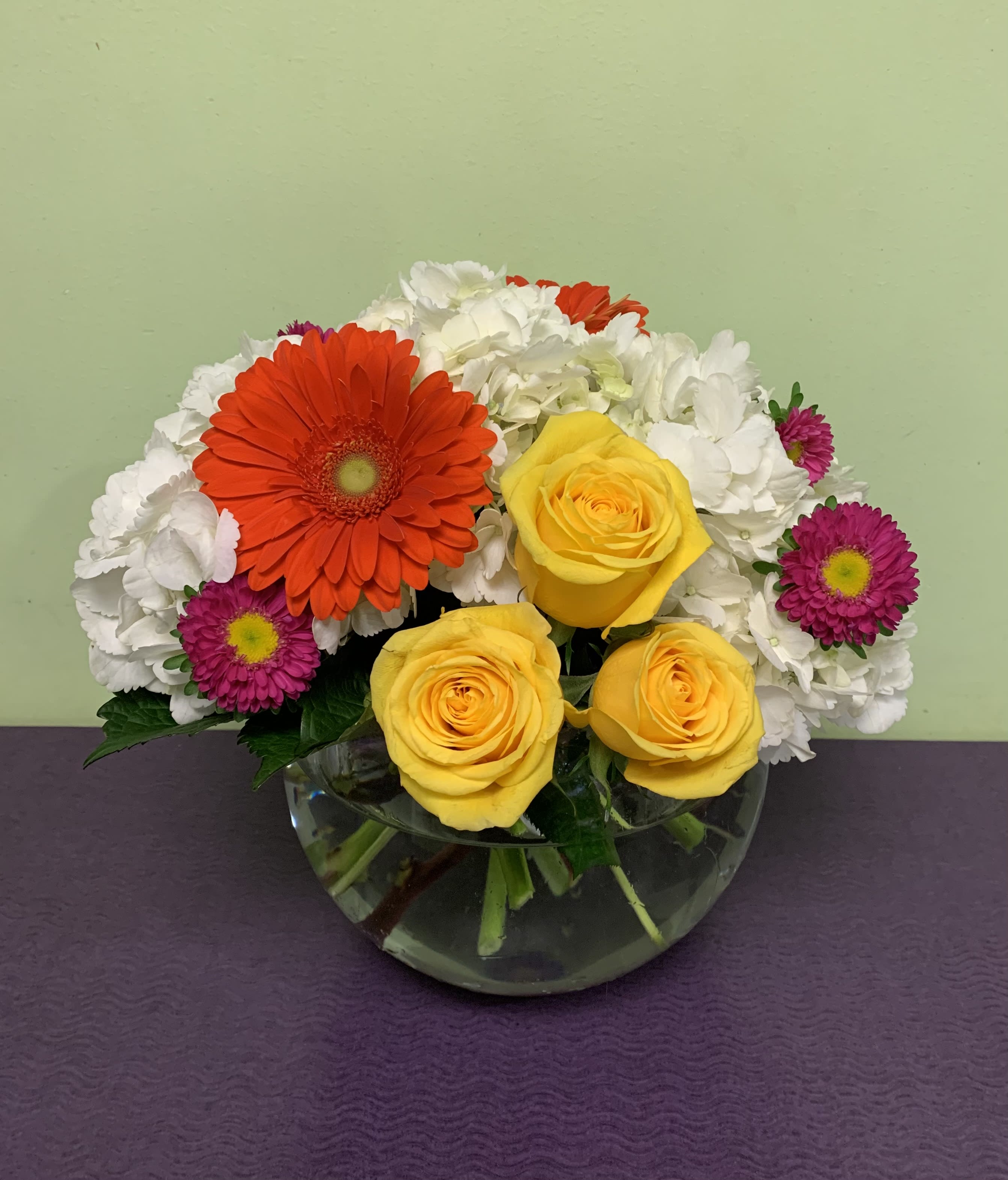 Mixed bouquet of yellow roses, orange gerbera daisies, and white blooms in a glass bowl vase