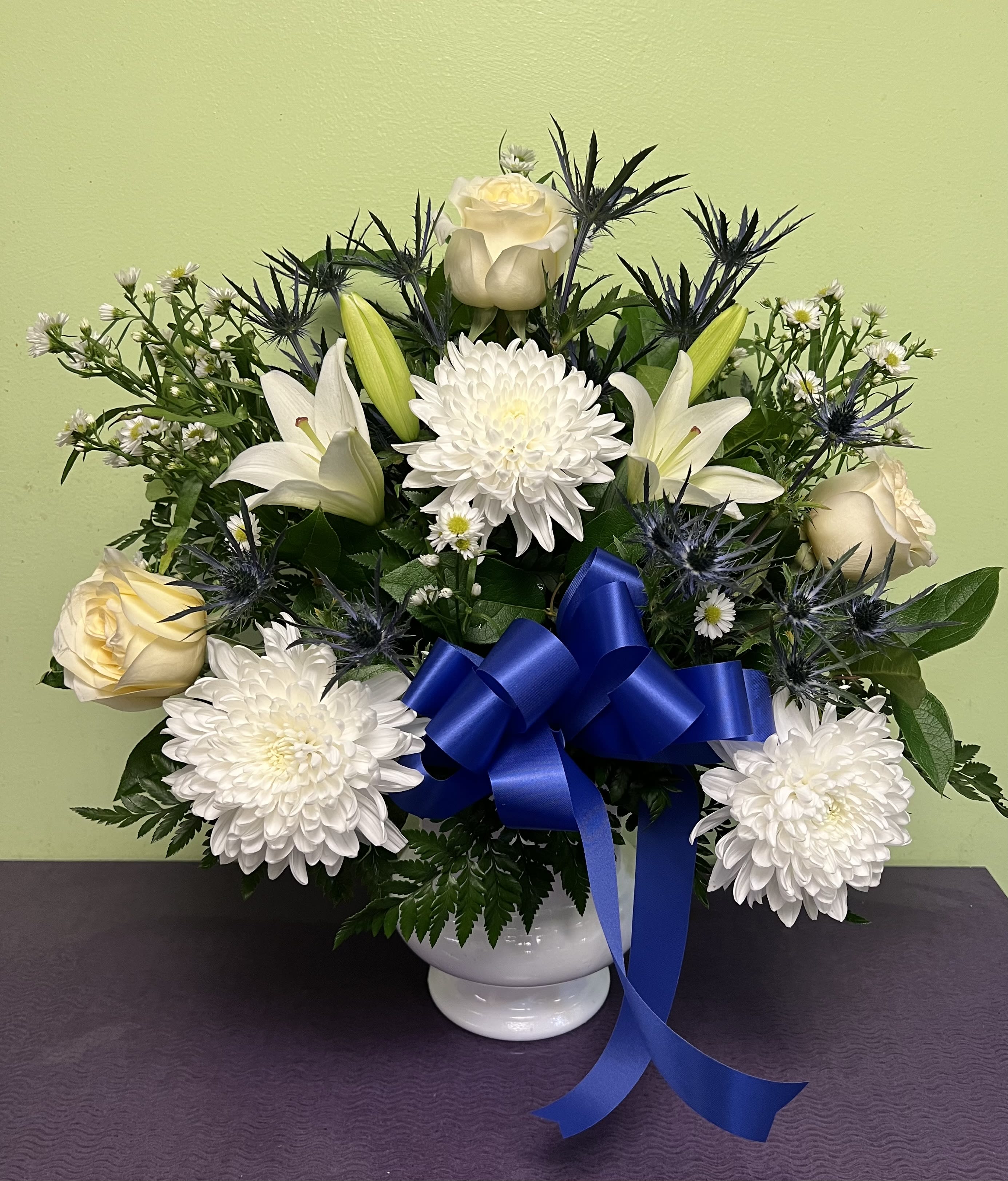 White roses and lilies arranged with chrysanthemums in a white vase with a blue ribbon