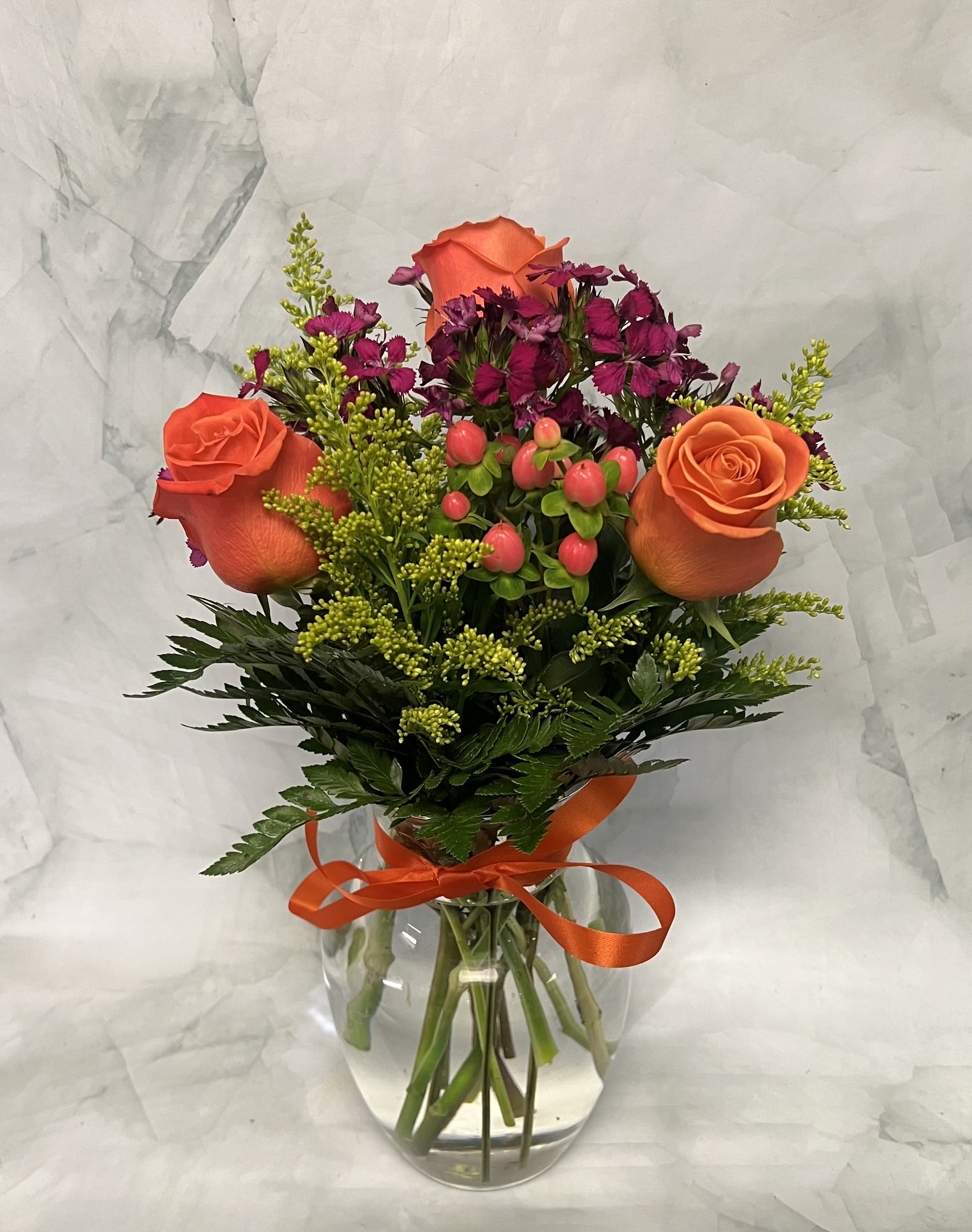 Orange roses and magenta flowers in a clear glass vase with an orange ribbon