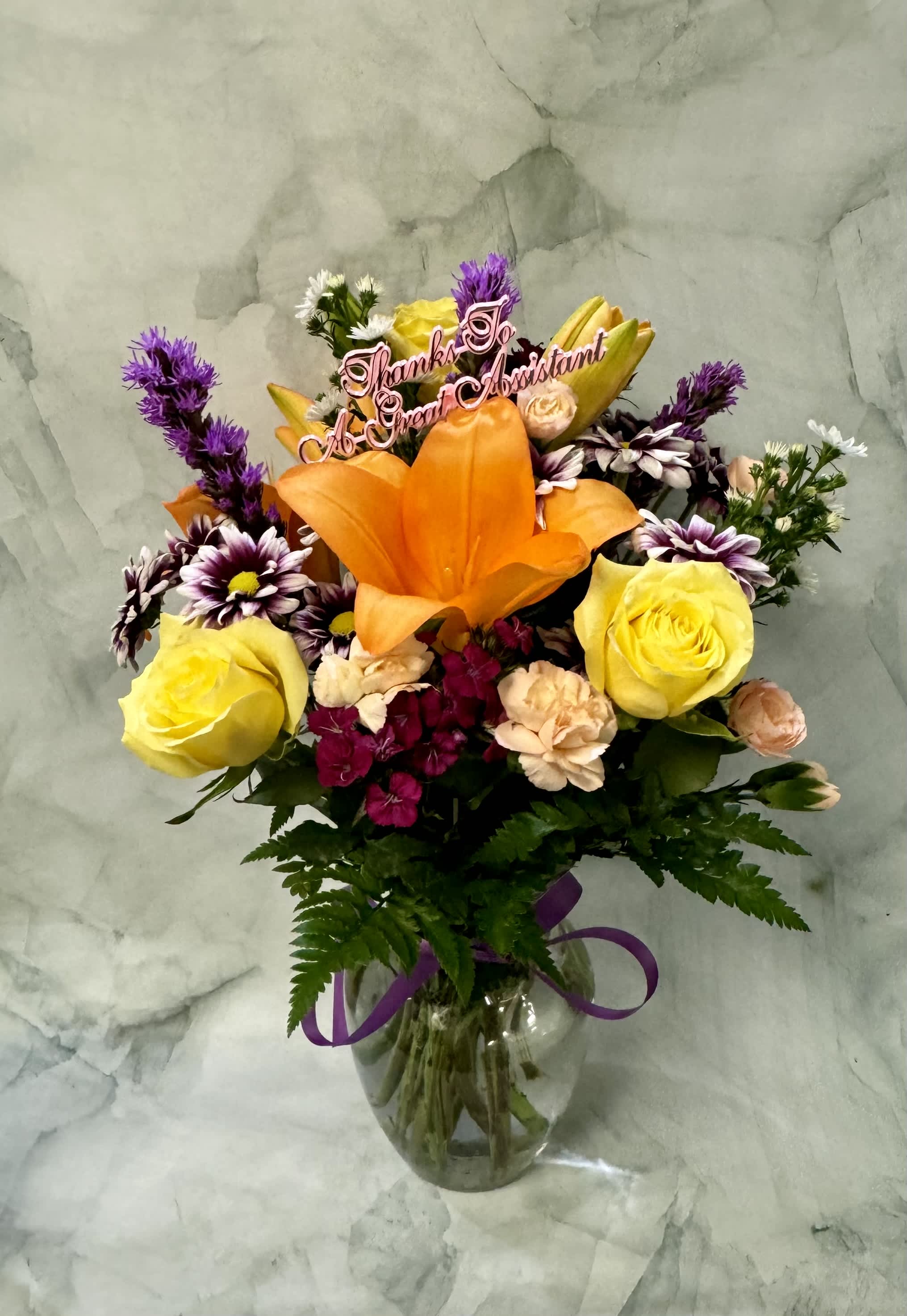 Mixed bouquet with orange lily, yellow roses, and daisies in a glass vase with a thank-you pick.