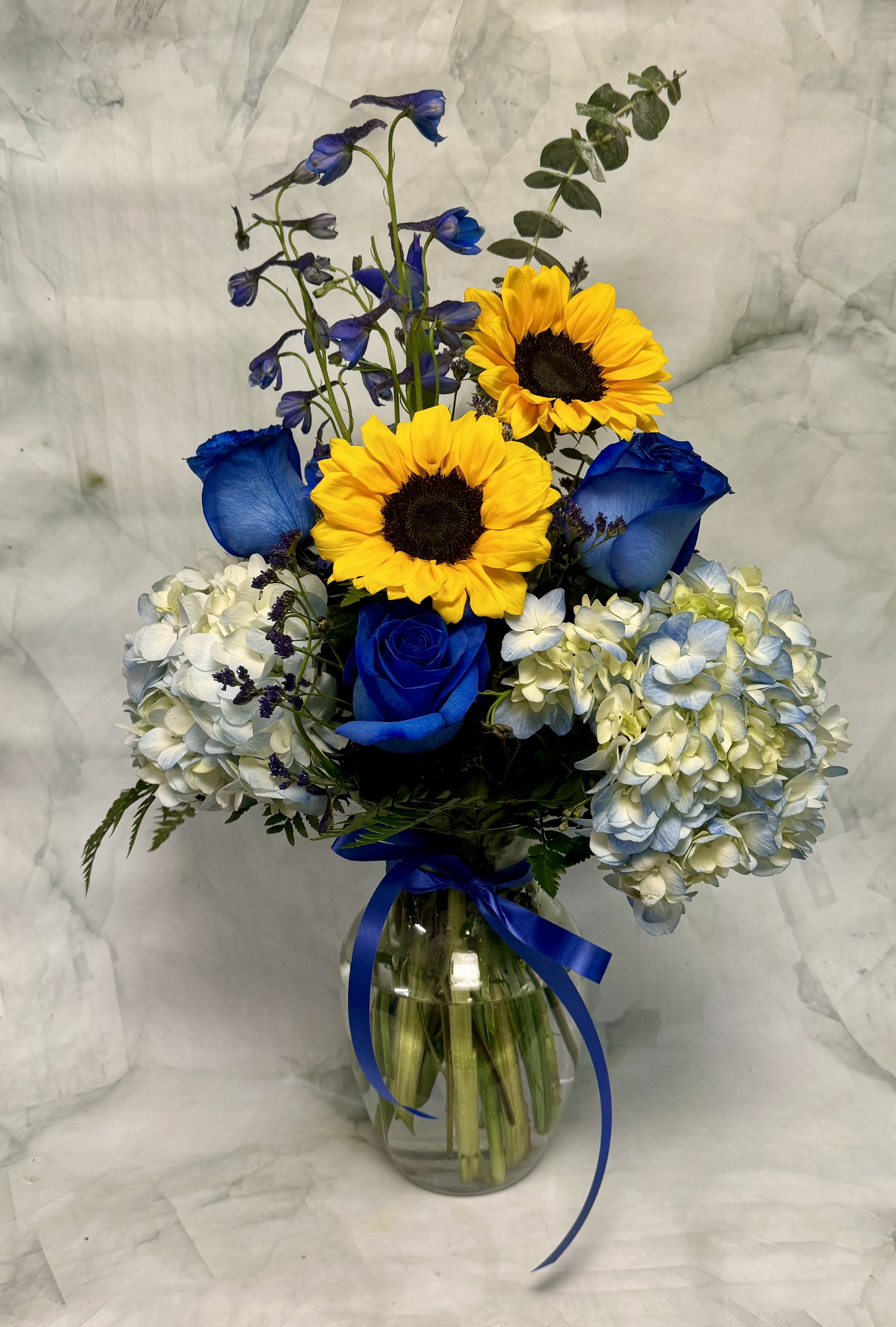 Blue roses and sunflowers in a glass vase with hydrangeas and a ribbon