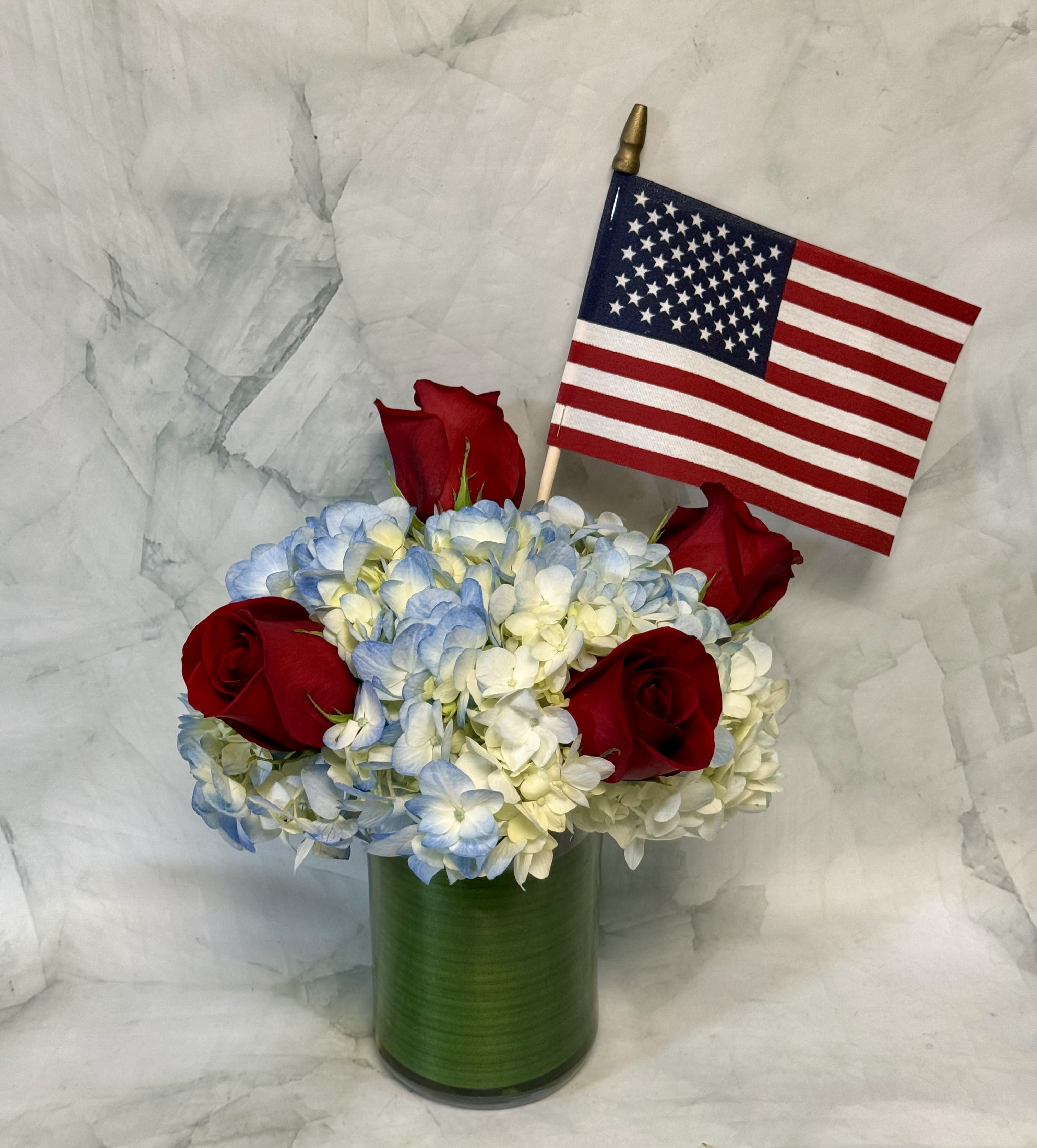 Red roses and blue-white hydrangeas in a green vase with a small U.S. flag