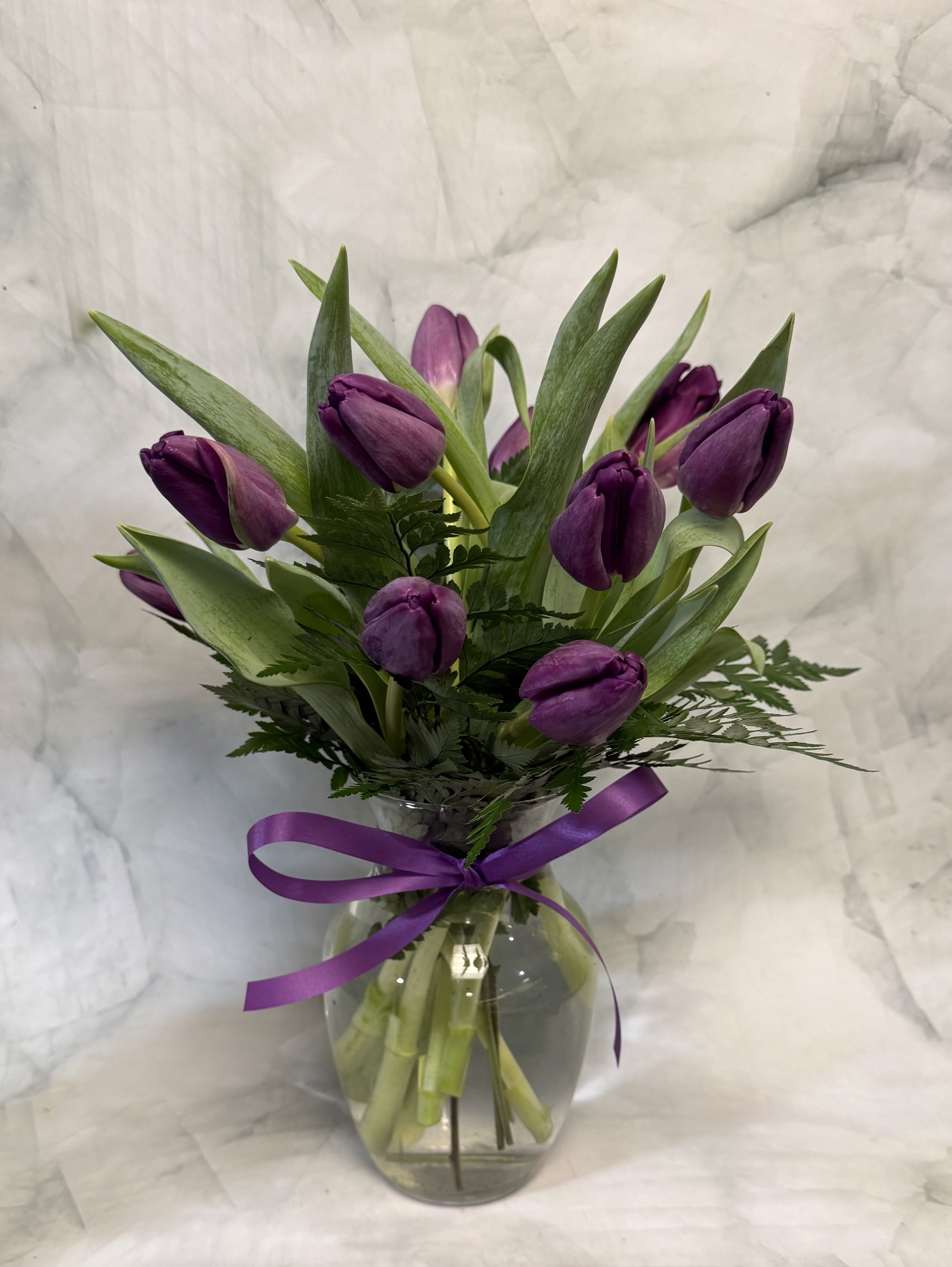 Purple tulips arranged in a clear glass vase with a purple ribbon.