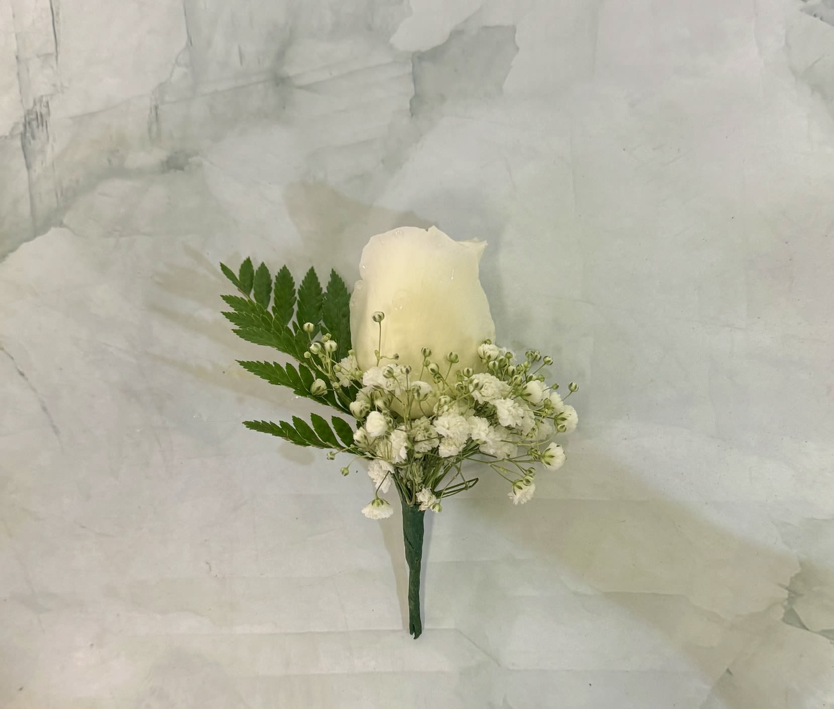 Simple White Rose Boutonnière - A single white rose boutonnière that reads as quietly elegant-the perfect finishing touch for a wedding, formal event, or sympathy service. This boutonnière is designed for the person you want to look composed and honored: the white rose and restrained green palette convey purity and respect while keeping the look classic and wearable.  The arrangement centers one fresh white rose, surrounded by tiny white filler blossoms and a delicate fern frond for contrast and texture. The stem is neatly wrapped in green floral tape for a clean, formal finish; the compact scale sits comfortably on a lapel without overpowering the wearer. Colors: white and green. Flowers: roses plus small filler blooms.  Ideal for grooms, fathers, or anyone you want to mark with quiet elegance, this boutonnière pairs easily with traditional suits and muted color palettes. It arrives ready to pin and wear-handle gently, keep cool, and place in water briefly before service if needed to ensure peak freshness.