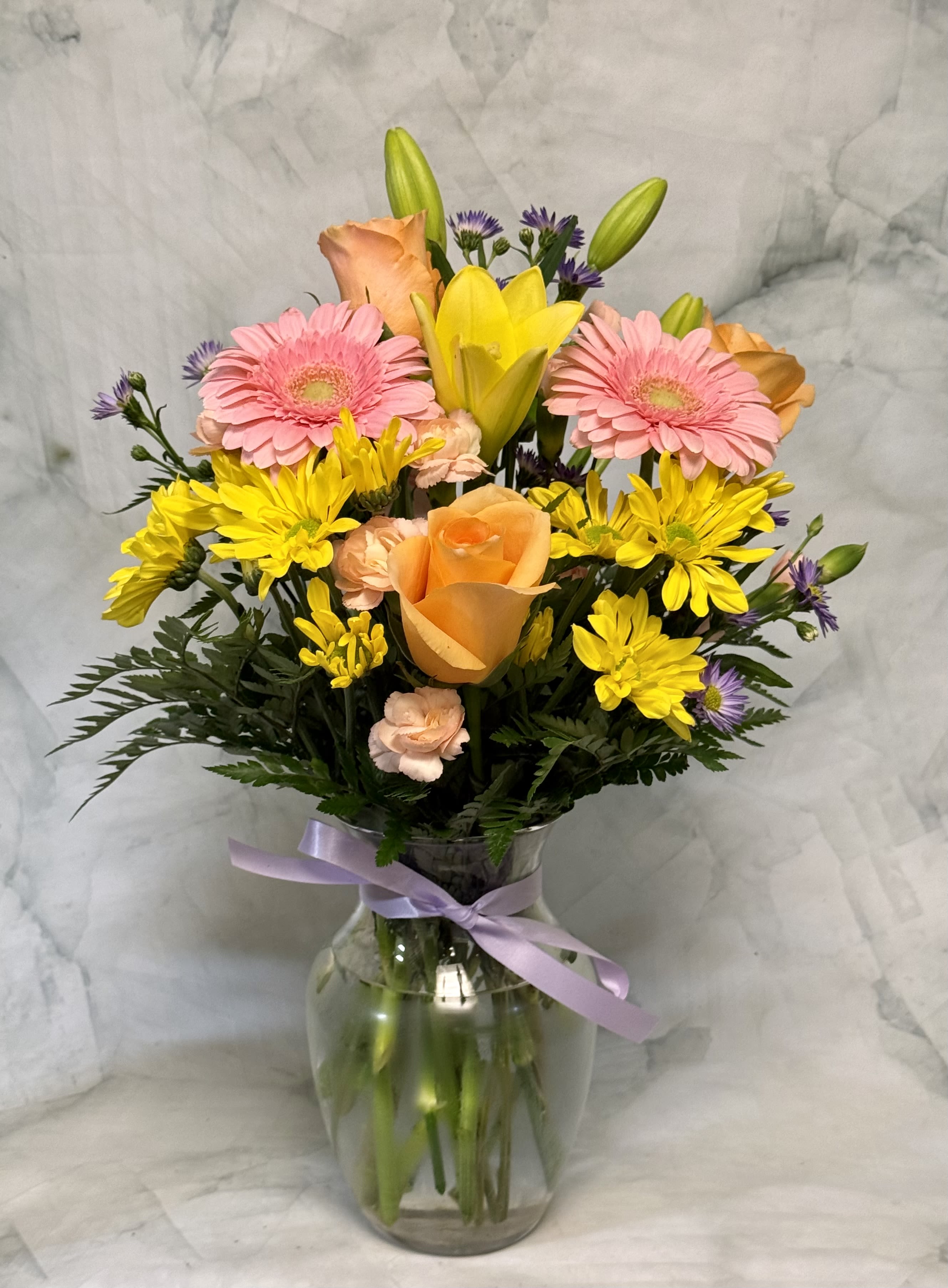 Spectacular Summer - A burst of sunlit color greets the recipient with bright yellow daisies and Asiatic lilies paired with pink gerbera daisies and soft peach roses-a summer flower arrangement that feels both celebratory and effortlessly cheerful. Ideal for summer parties, a thank-you gift, or a graduation bouquet, this arrangement is designed to make the person you're thinking of feel noticed and appreciated.  The hand-tied mix includes carnations, dianthus, chrysanthemums, asters and other seasonal filler blooms in shades of purple and pastel to add contrast and texture. Stems are arranged in a clear glass vase and finished with a lavender ribbon for a simple, classic presentation that showcases the blooms. The composition reads full and lively without feeling fussy, perfect for sending congratulations, saying thanks, or brightening someone's day; vase-ready on arrival and easy to display wherever they'll enjoy it most.